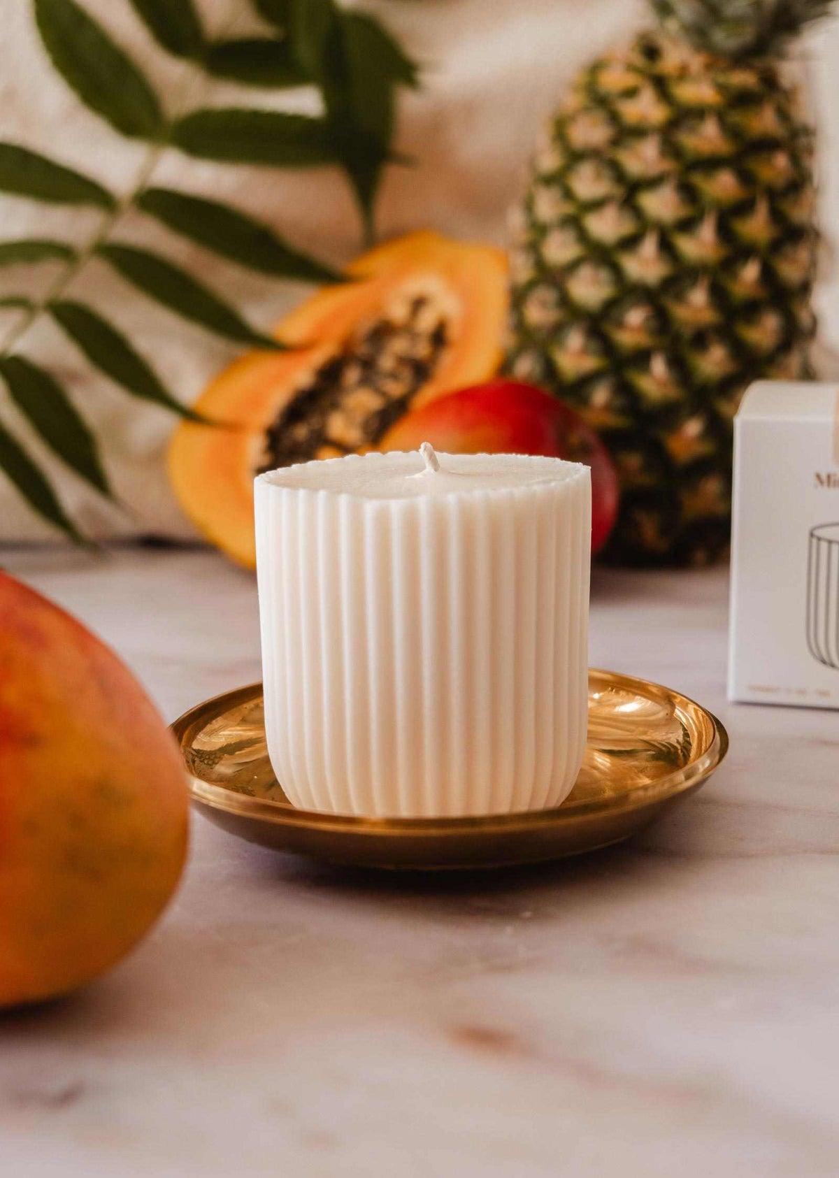 The Candle Refill Coco Mango by Mimi & August, a white ribbed tropical candle, sits on a gold plate with mango, papaya, pineapple, and green leaves in the background.