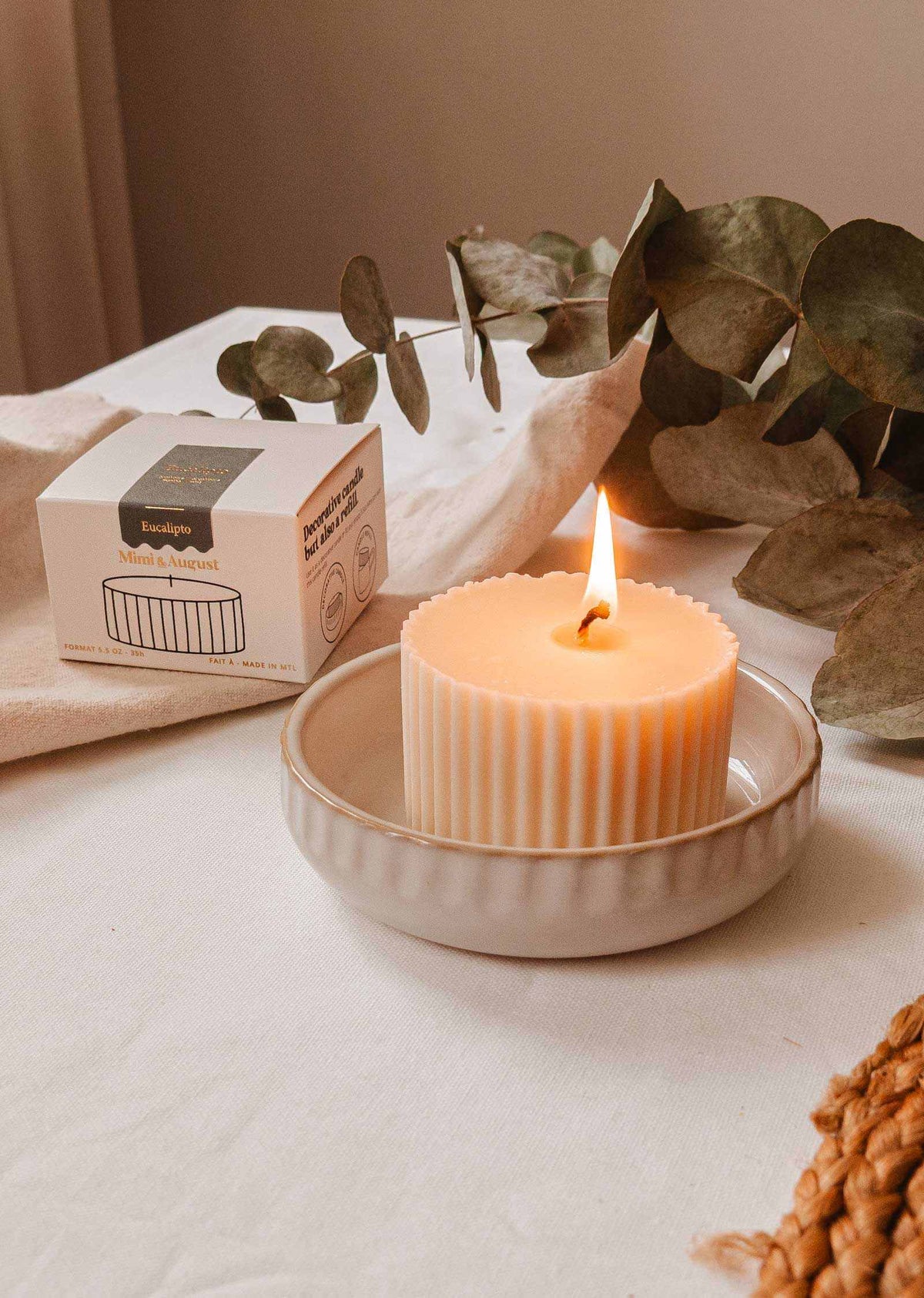 The Candle Refill - Eucalipto by Mimi & August sits gracefully lit in a ceramic dish alongside its eco-friendly box and lush eucalyptus leaves, elegantly arranged on a cloth-covered surface.