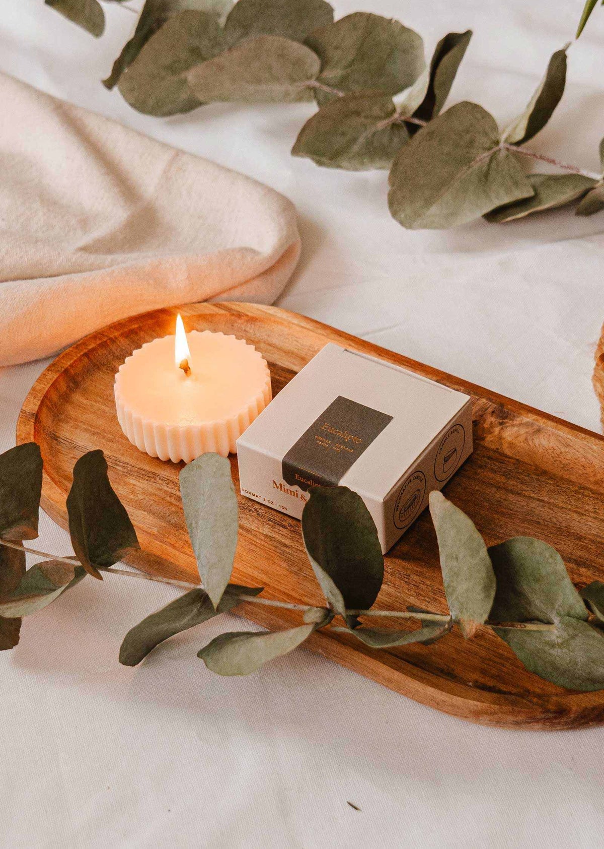 The Candle Refill - Eucalipto by Mimi & August sits elegantly on a wooden tray with a box, surrounded by lush eucalyptus leaves on a white surface, offering an eco-friendly touch.