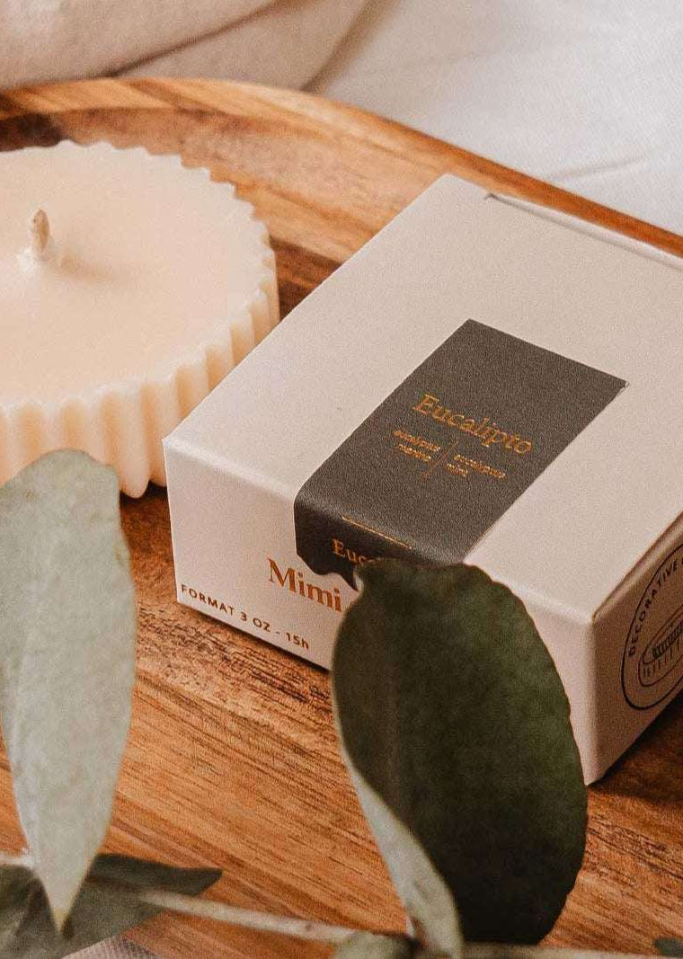 A Candle Refill - Eucalipto by Mimi & August and a boxed item sit on a wooden tray, surrounded by eucalyptus leaves on a white surface, creating an eco-friendly centerpiece.