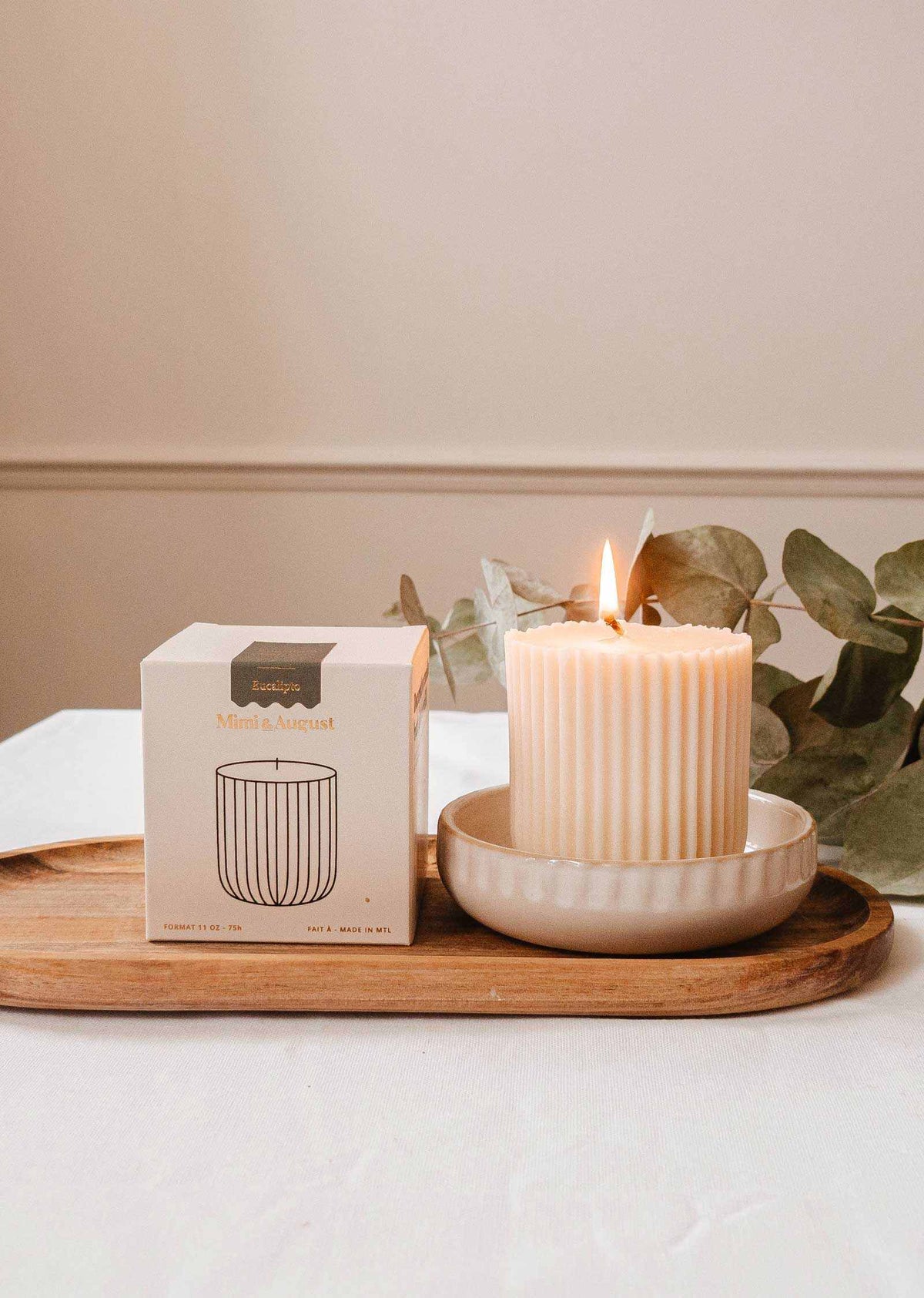 The Candle Refill Eucalipto by Mimi & August glows in a ceramic dish atop a wooden tray near its box, with eucalyptus branches in the background.