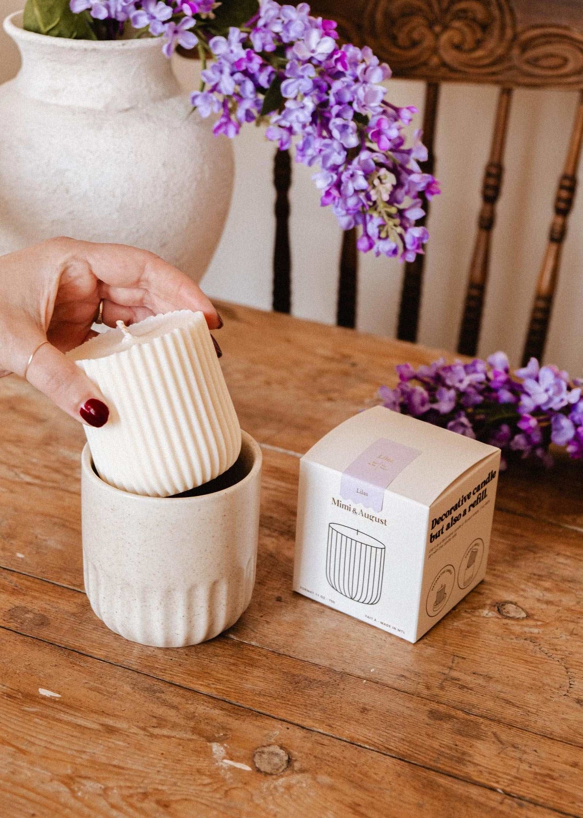 A hand places the Mimi & August Candle Refill Lilac, a ribbed white soy wax candle, into a ceramic holder beside its box, with purple flowers suggesting lilac fragrance and a wooden chair in the background.