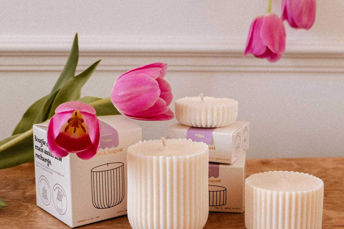 Three Mimi & August Candle Refill - Tulipa soy wax candles with ribbed white design sit in their boxes on a wooden surface, surrounded by pink tulips and set against a white wall.