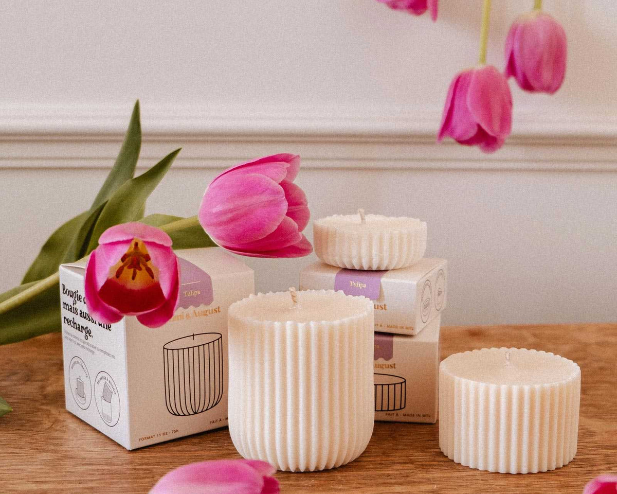 Three Mimi & August Candle Refill - Tulipa soy wax candles with ribbed white design sit in their boxes on a wooden surface, surrounded by pink tulips and set against a white wall.