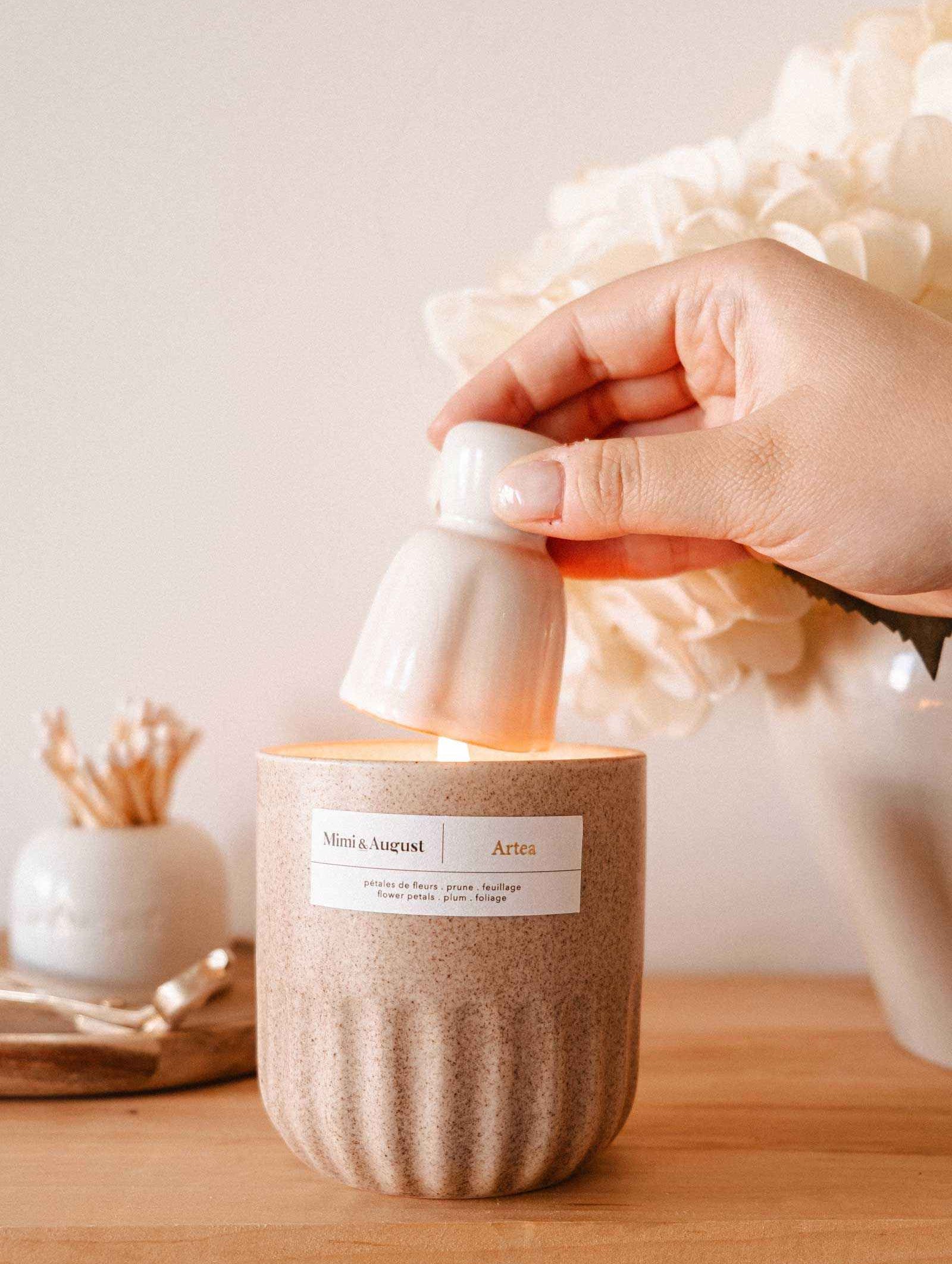 A hand uses the Mimi & August Candle Snuffer to extinguish a lit candle in a beige container on a wooden surface, with decorative candle accessories in the background.