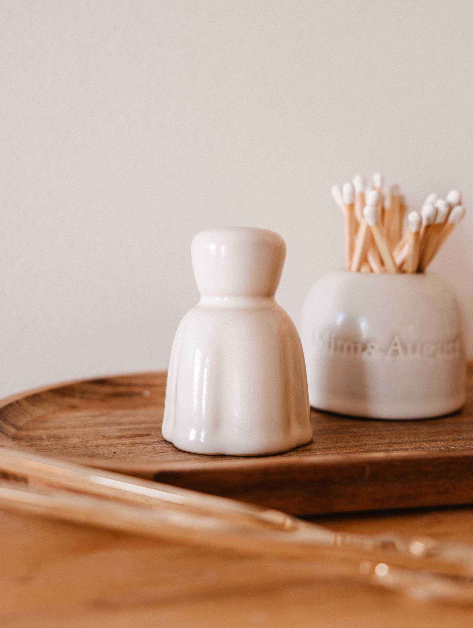 The Mimi & August Candle Snuffer, a small white ceramic piece, sits on a wooden tray beside a jar of cotton swabs, adding an elegant touch to your candle accessories collection.