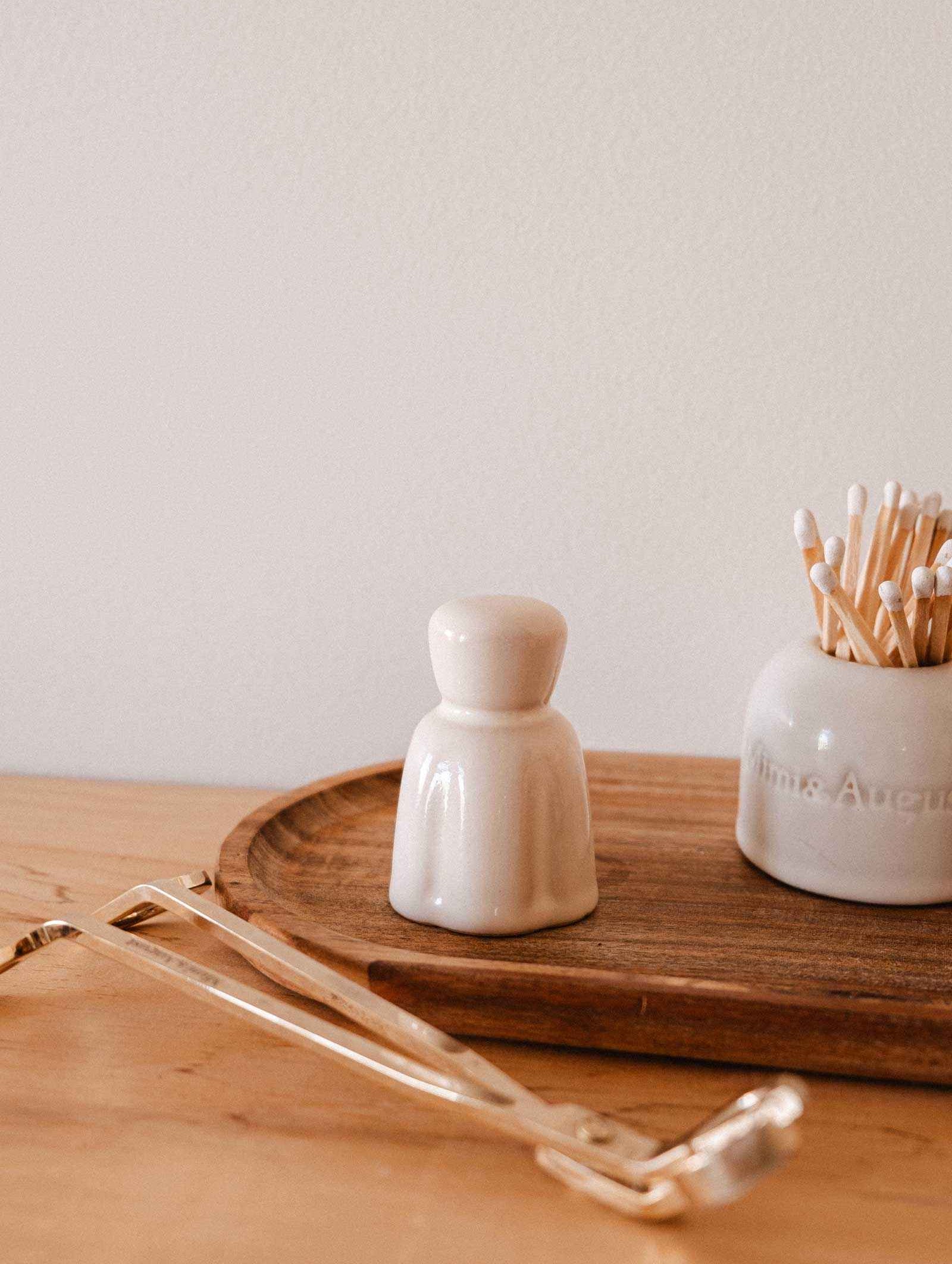 A Mimi & August Candle Snuffer rests on a light wood surface beside a small ceramic container, a jar of cotton swabs, and other candle accessories.