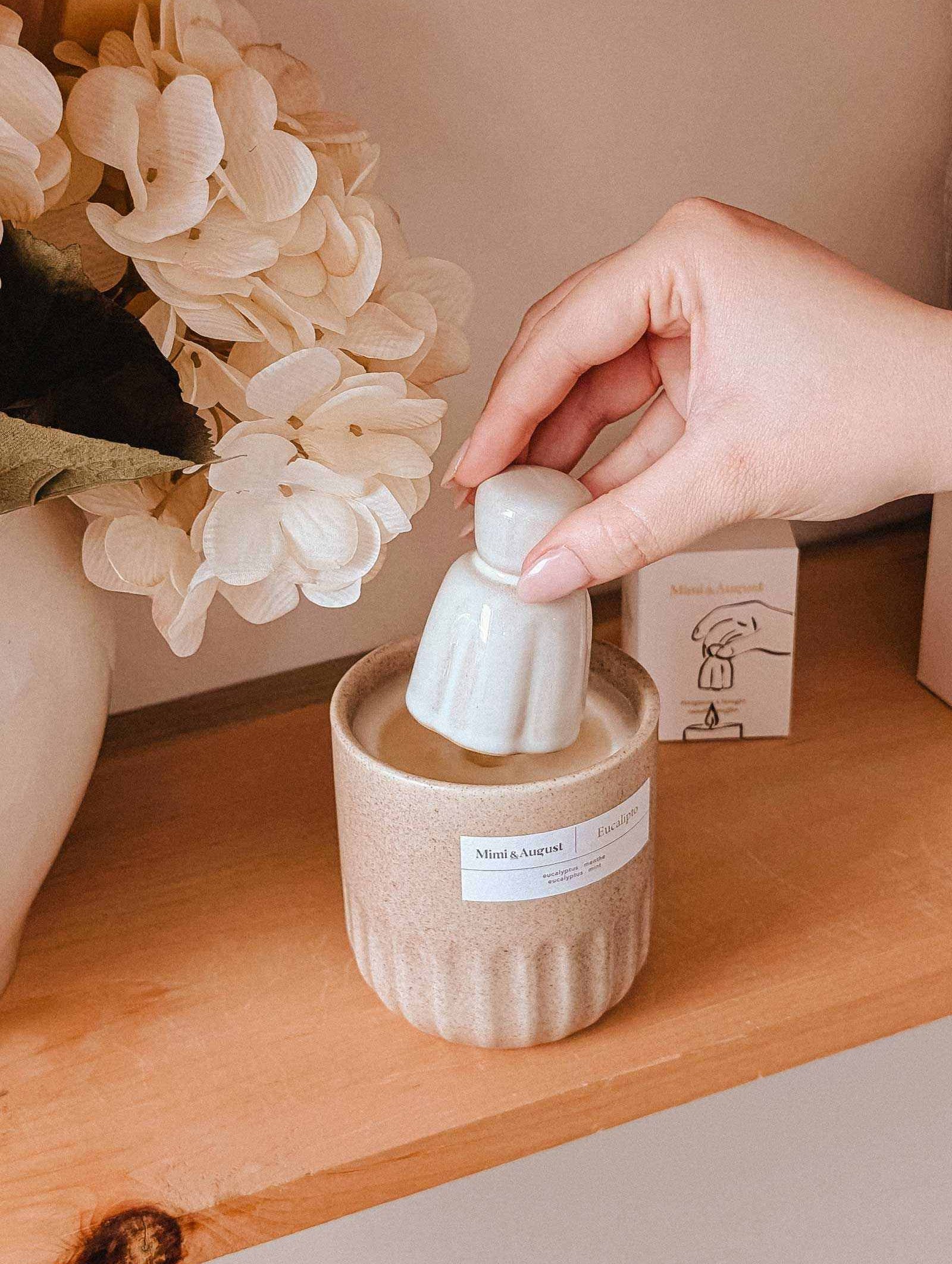 A hand sets the Mimi & August Candle Snuffer into a candle jar on a wooden shelf beside a vase of white flowers, bringing charm to essential candle accessories.