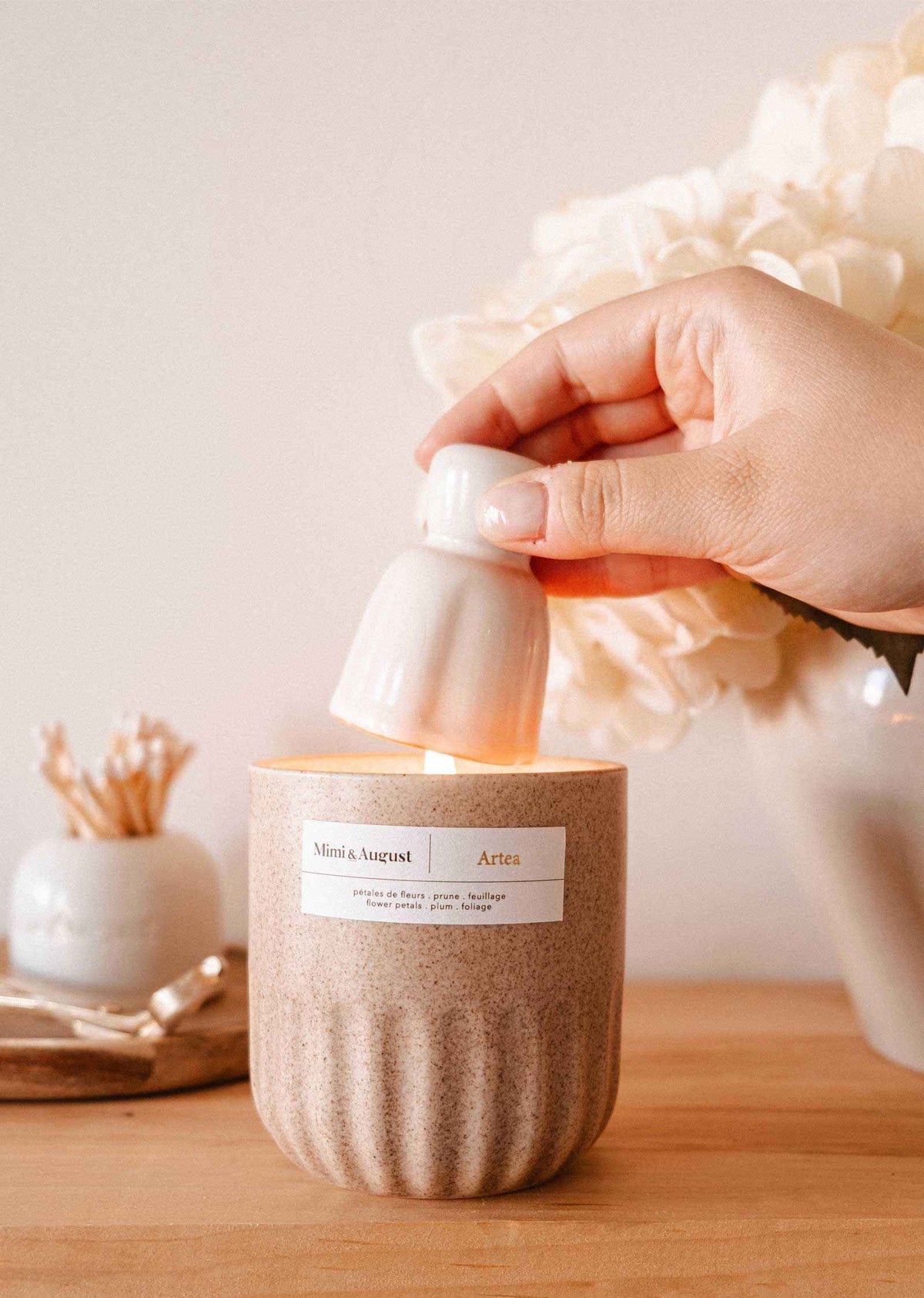 A hand uses the Mimi & August Candle Snuffer to extinguish a lit candle in a beige container on a wooden surface, with decorative candle accessories in the background.