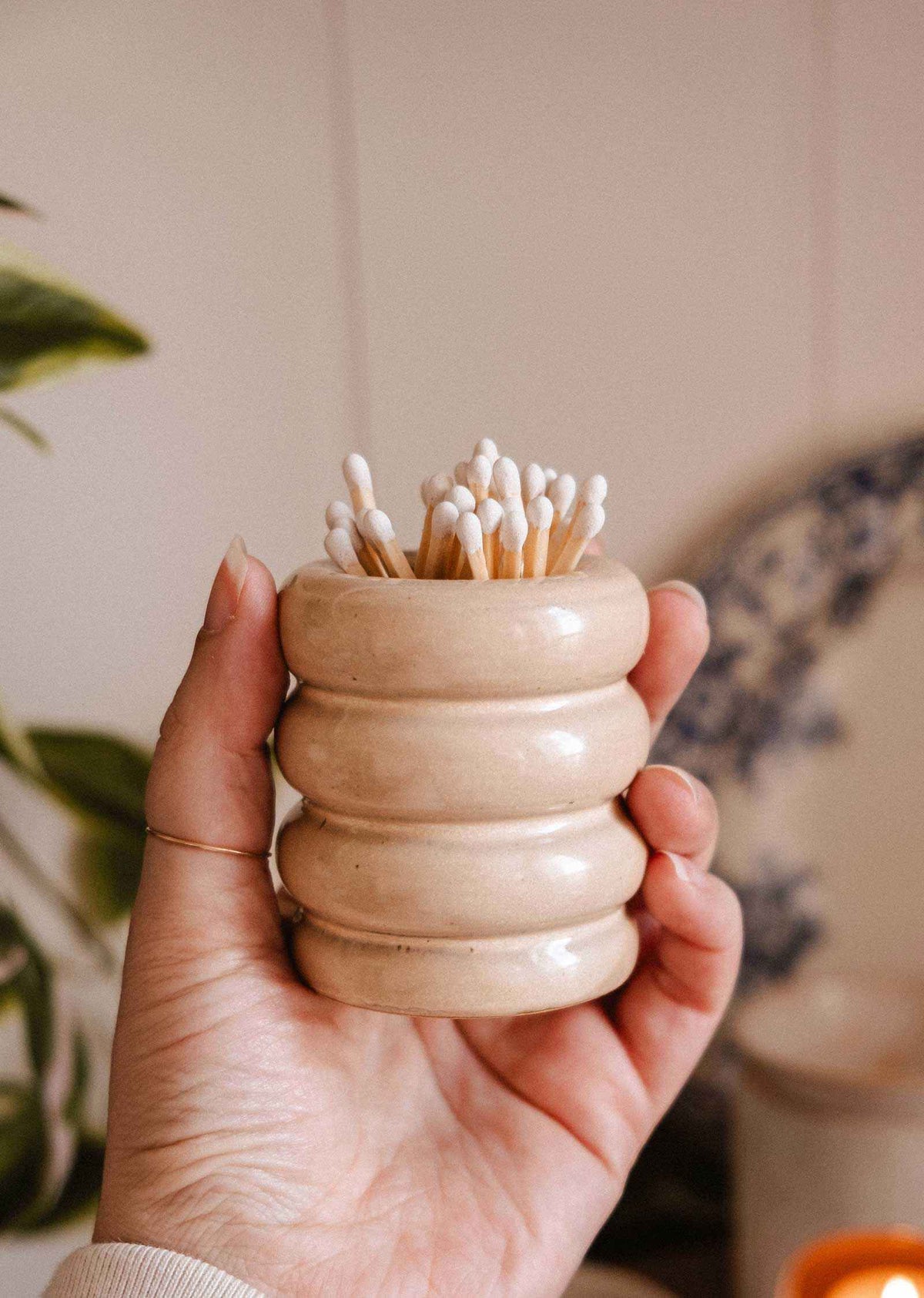 A hand holds the Mimi & August Ceramic Match Pot in beige, featuring a ribbed design and filled with cotton swabs. A blurred plant and plate appear in the background, highlighting the charm of this decorative candle accessory.