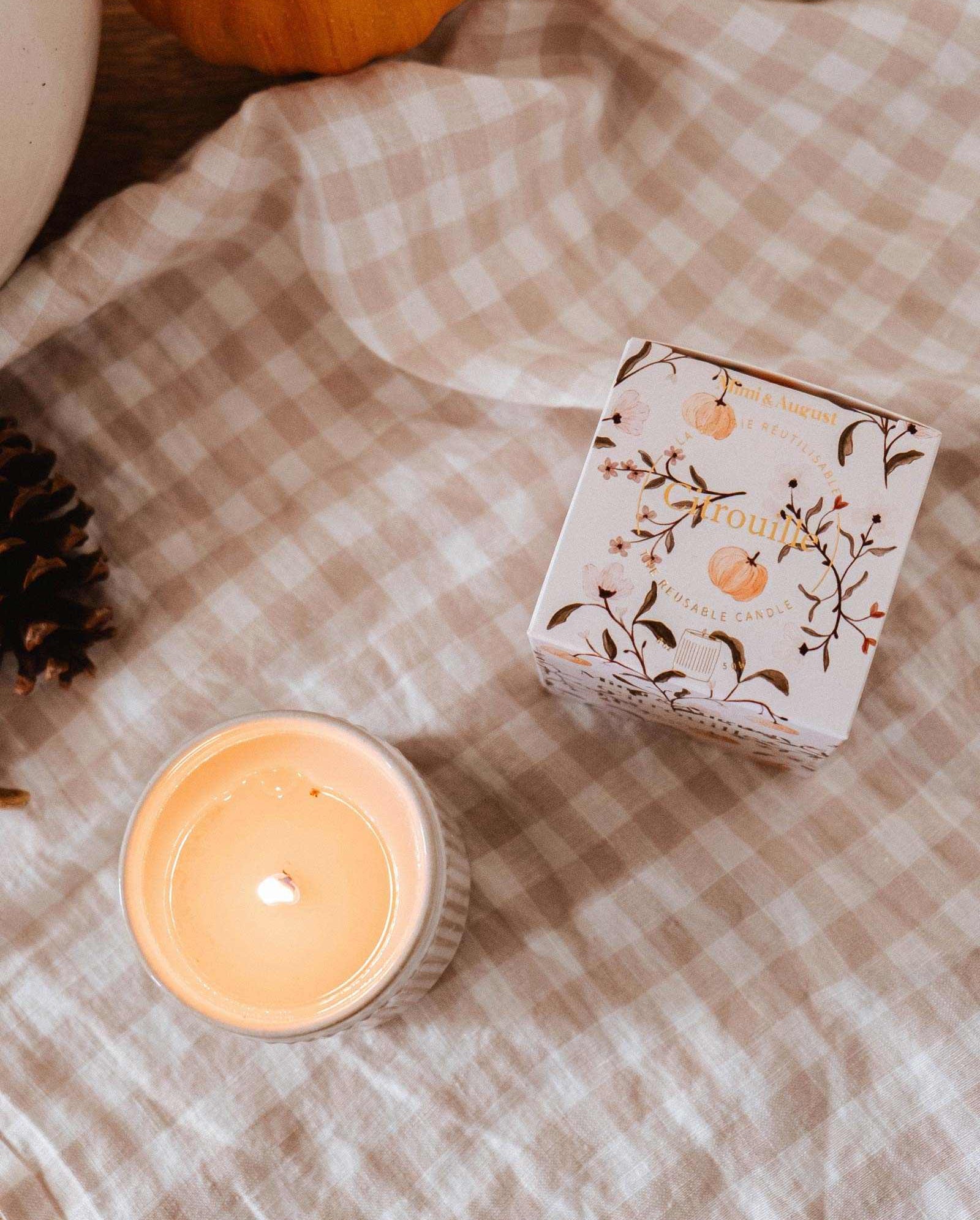 A lit Citrouille - Reusable Candle by Mimi & August in a white jar sits next to its decorative box on a beige checkered cloth, with a pinecone and pumpkin partly visible.