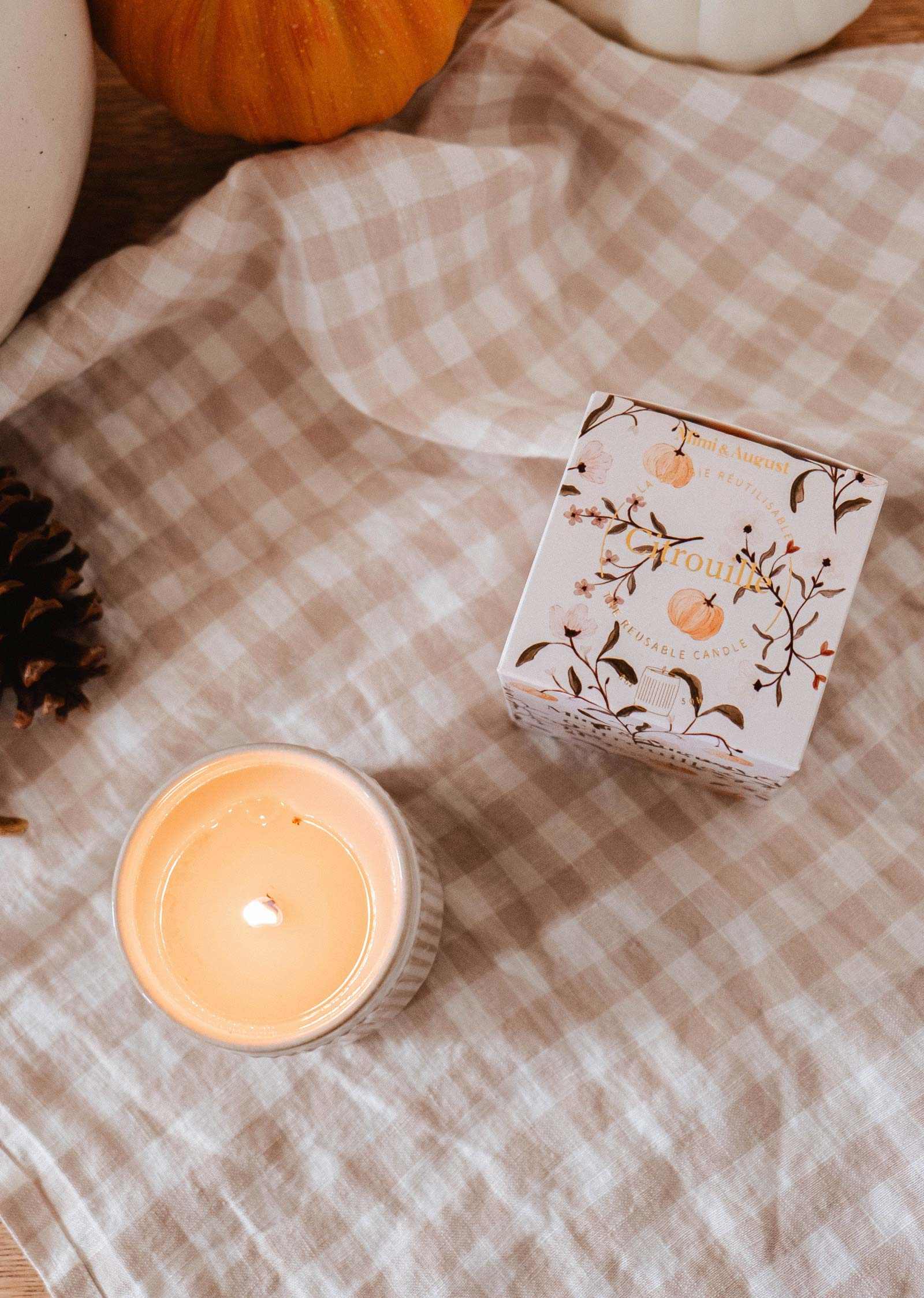 A lit Citrouille - Reusable Candle by Mimi & August in a white jar sits next to its decorative box on a beige checkered cloth, with a pinecone and pumpkin partly visible.