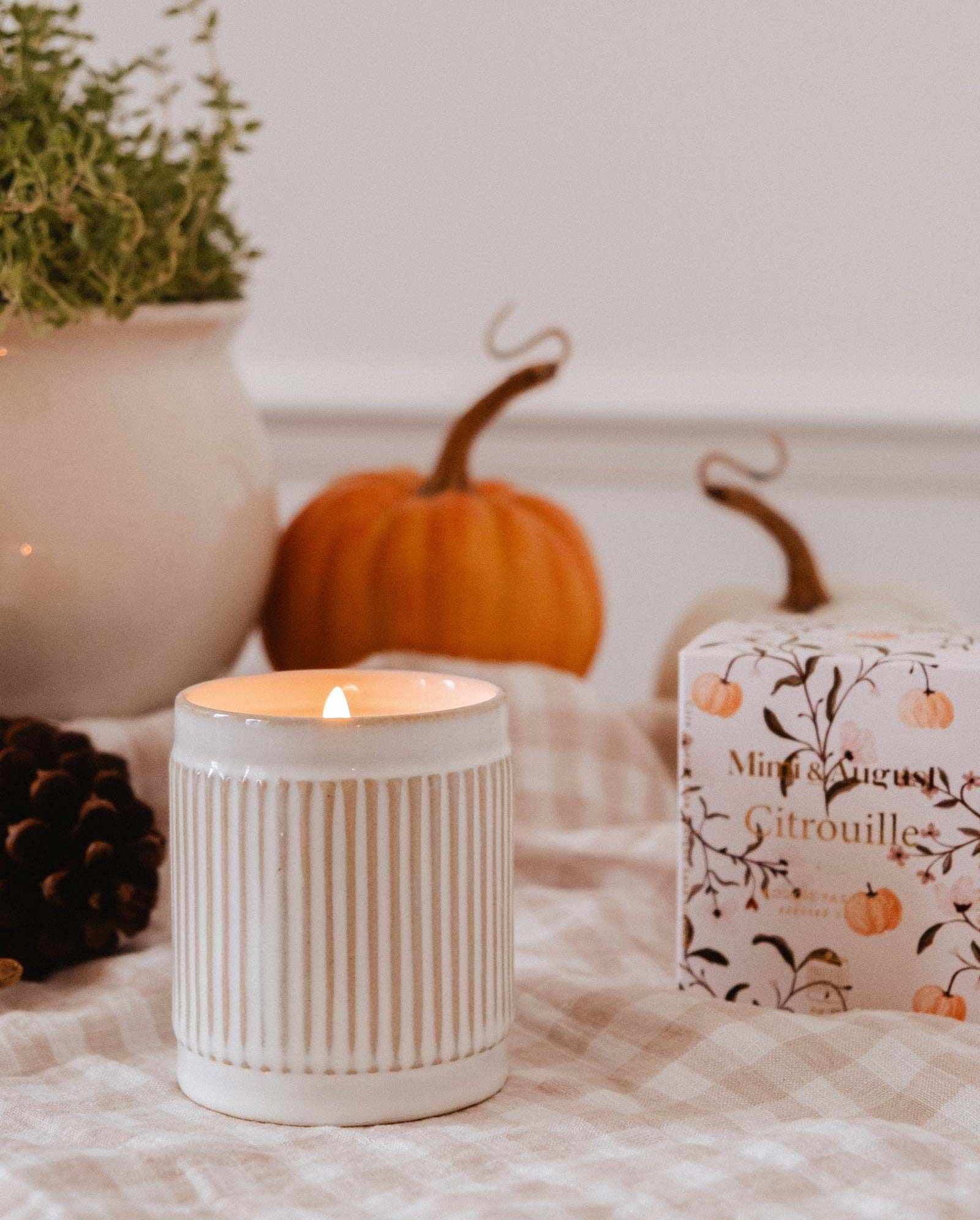 A lit Mimi & August Citrouille Reusable Candle, a pinecone, small pumpkins, a potted plant, and a decorative box are arranged on a checkered cloth.