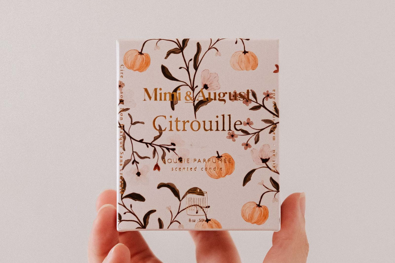 A hand holds a small "Citrouille - Reusable Candle" box from Mimi & August, decorated with orange pumpkins and branches, revealing a charming pumpkin spice candle inside.