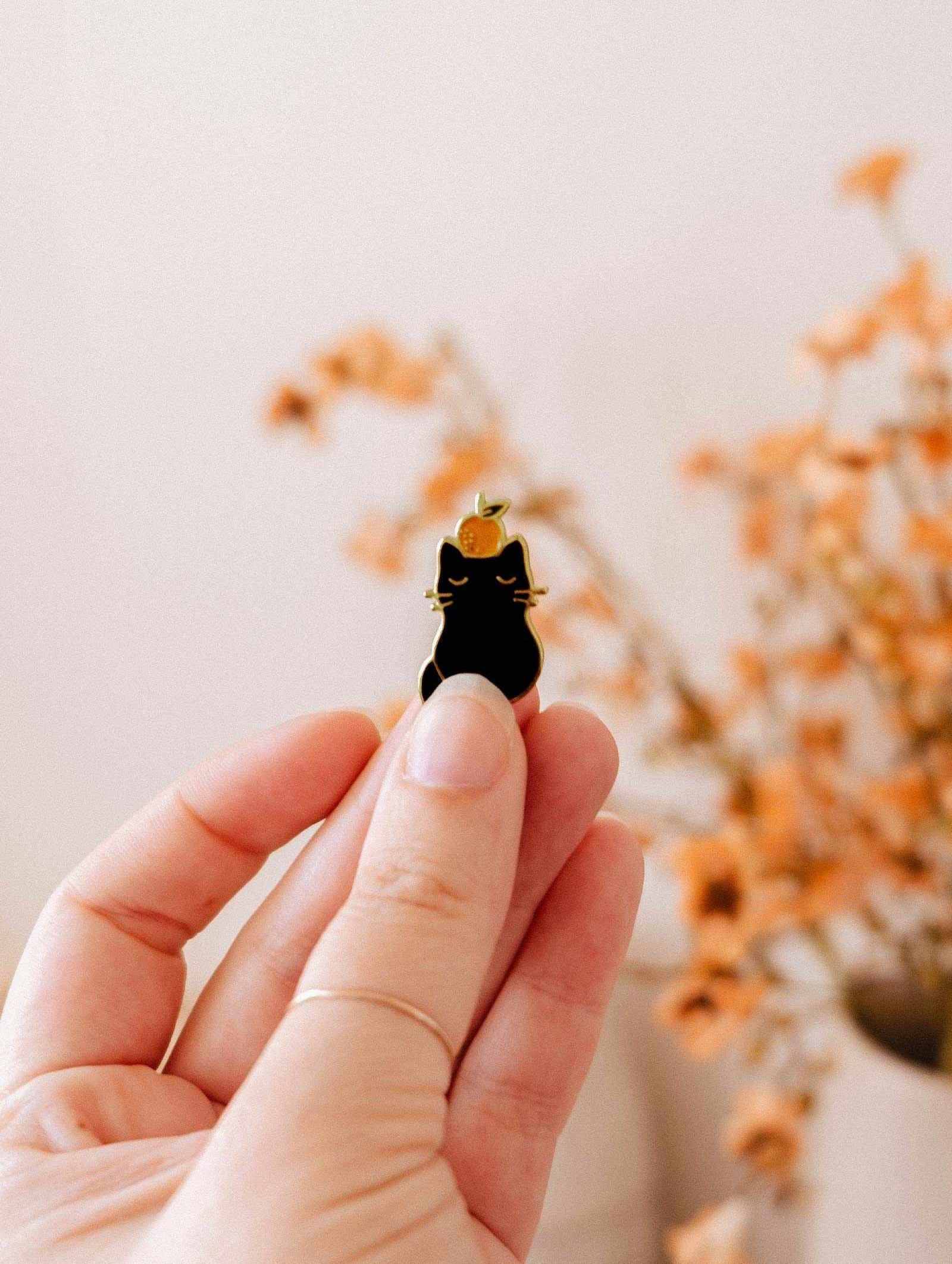 A hand holds the Clementine Cat Enamel Pin by Mimi & August, a quirky small black cat accessory, with a blurred background featuring orange dried flowers.