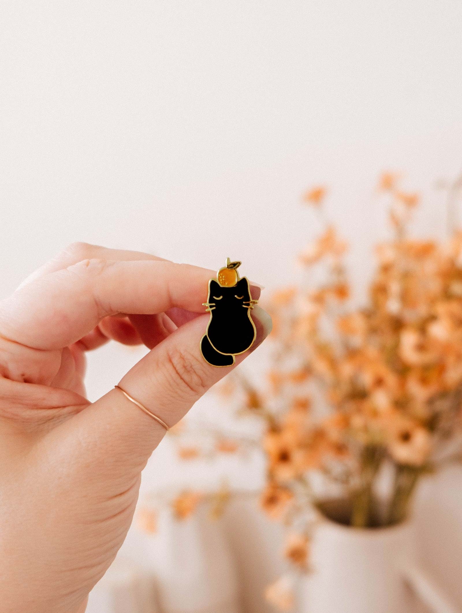 A hand holds the Mimi & August Clementine Cat Enamel Pin with gold outlines; a blurred vase of orange flowers appears in the background.