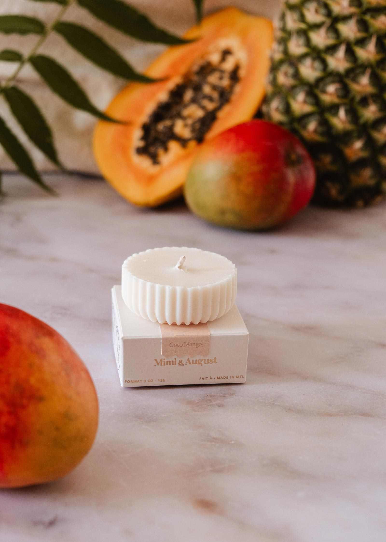 The Candle Refill Coco Mango by Mimi & August sits on a branded box, surrounded by mango, papaya, and pineapple, atop a marble surface.