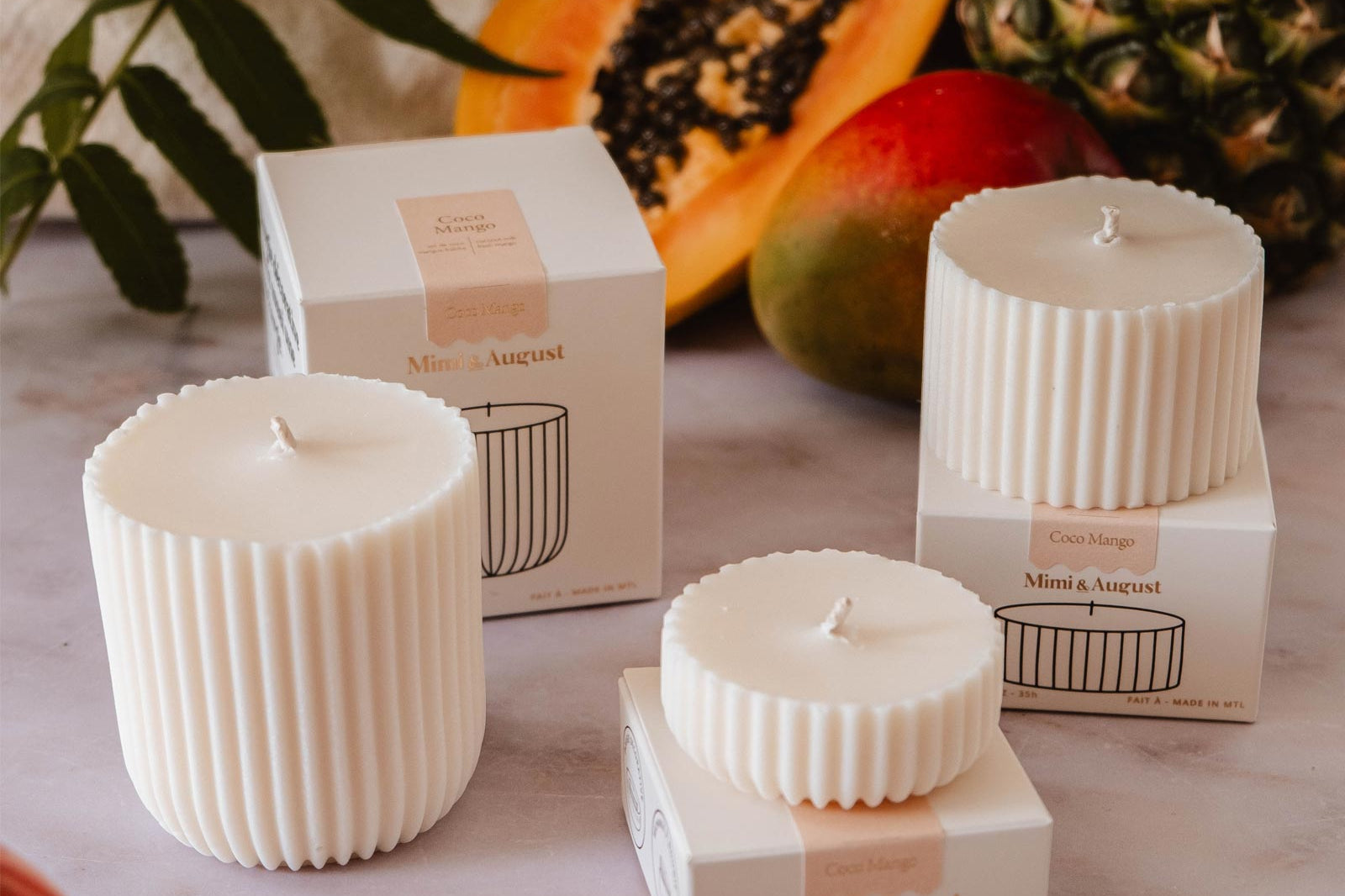 Three white ribbed candles on a marble surface, each placed near a matching box. Surrounding the candles are tropical fruits like papaya, pineapple, and mango, along with green foliage. Enjoy the ambiance with our eco-friendly Candle Refill - Coco Mango from Mimi & August for an exotic touch.