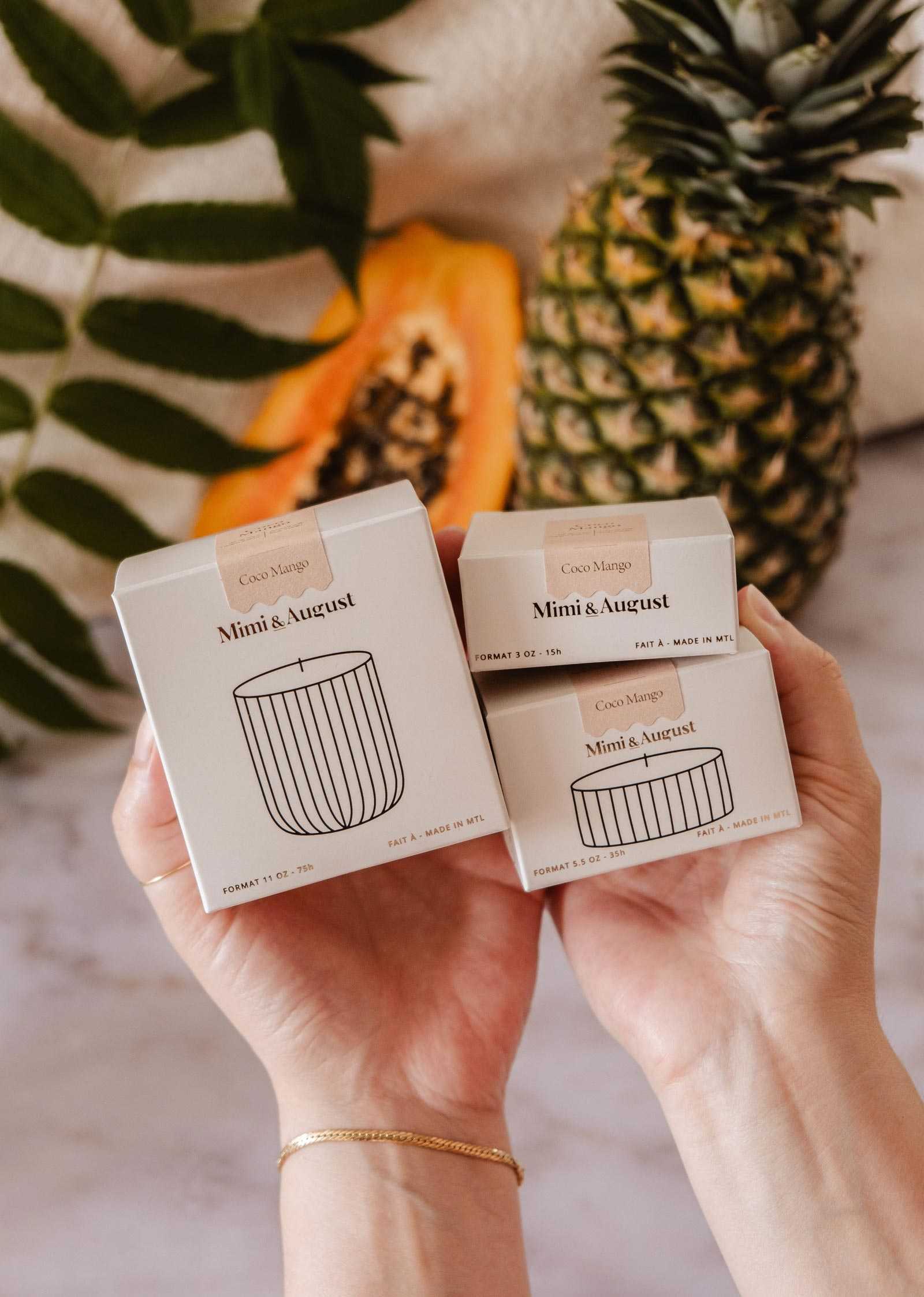 Hands hold three Mimi & August Candle Refill Coco Mango boxes, with pineapple, papaya, and green leaves in the background on marble, creating a fresh tropical vibe.