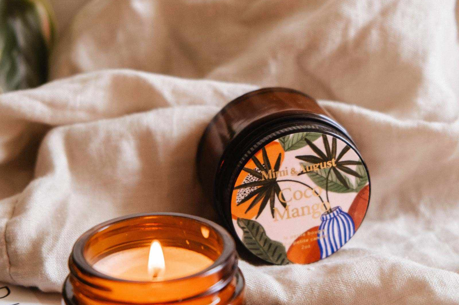 Une bougie de soya Coco Mango de 2 oz de Mimi & August allumée dans un pot en verre ambré emplit l'air d'un parfum tropical. La bougie réutilisable repose sur un livre à côté de son couvercle, avec un tissu beige et des feuilles vertes en arrière-plan.