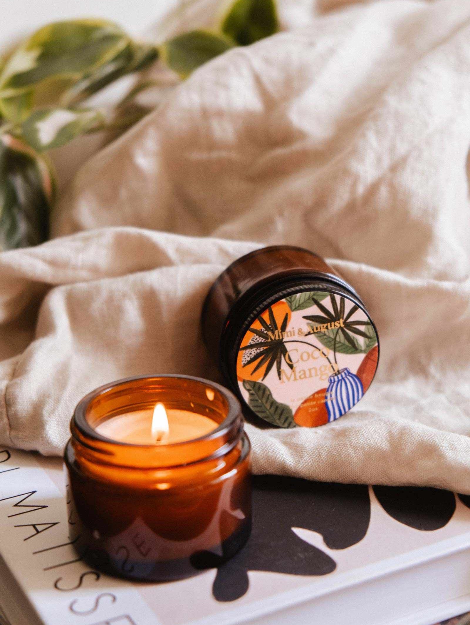 Une bougie de soya Coco Mango de 2 oz de Mimi & August allumée dans un pot en verre ambré emplit l'air d'un parfum tropical. La bougie réutilisable repose sur un livre à côté de son couvercle, avec un tissu beige et des feuilles vertes en arrière-plan.