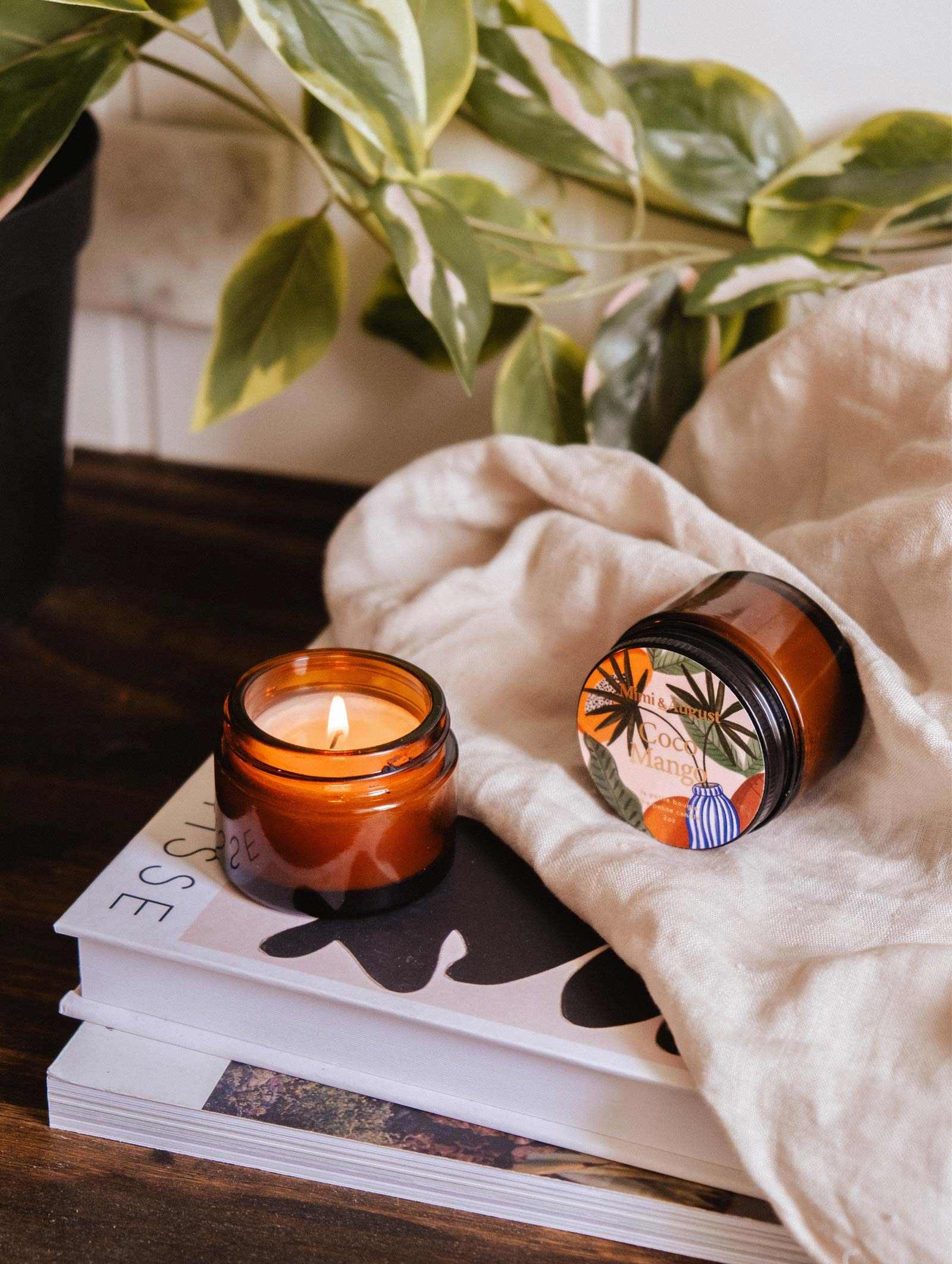 Une bougie de soya Coco Mango de 2 oz de Mimi & August brûle à côté d'un pot fermé, d'un tissu beige, d'une plante verte et de deux livres empilés sur une surface en bois.