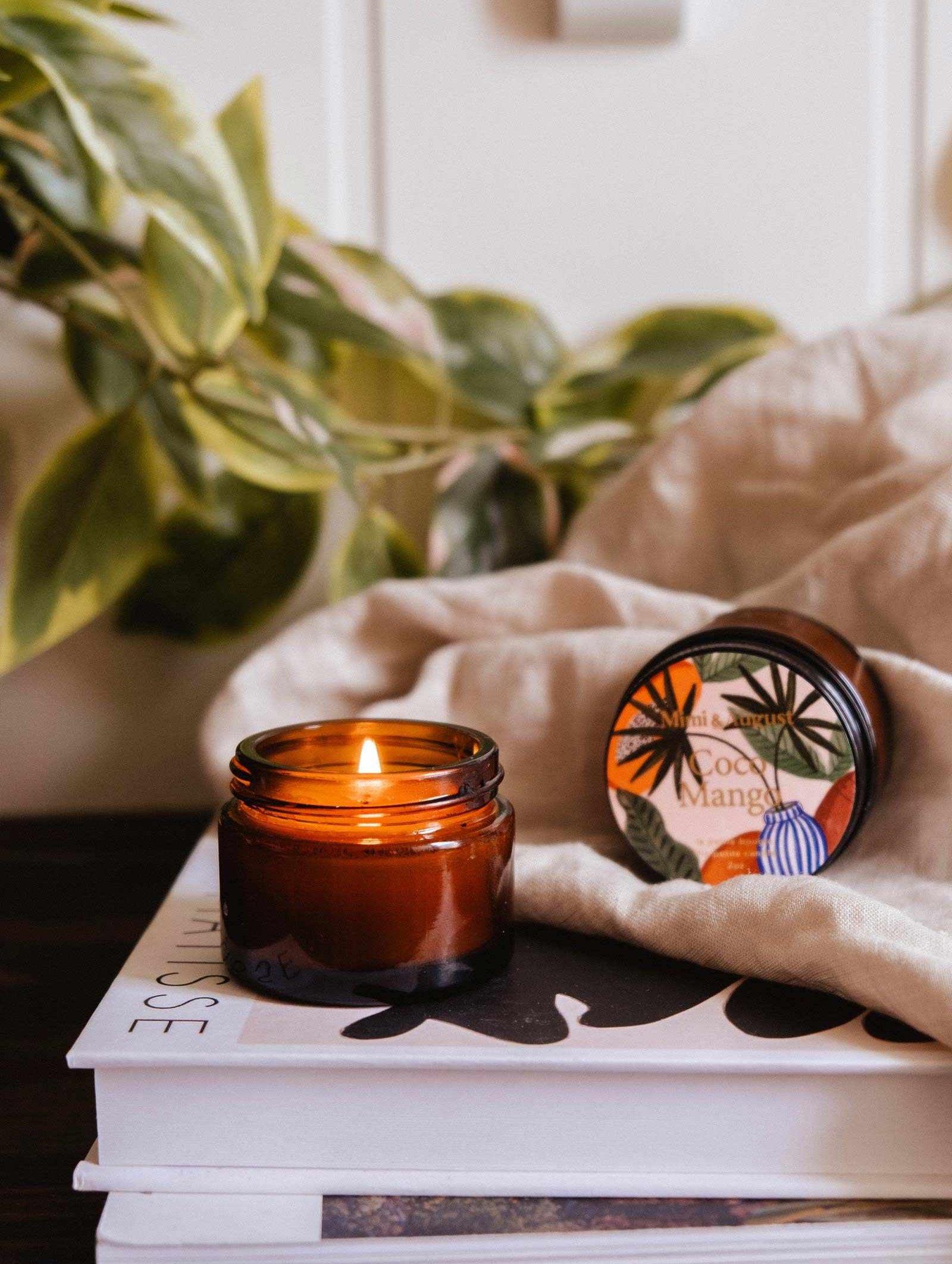 Une bougie de soya Coco Mango de 2 oz de Mimi & August allumée repose sur des livres empilés à côté d'un tissu, d'une plante et d'une bougie fermée étiquetée « Coco Mango », emplissant l'espace d'un chaleureux arôme tropical.