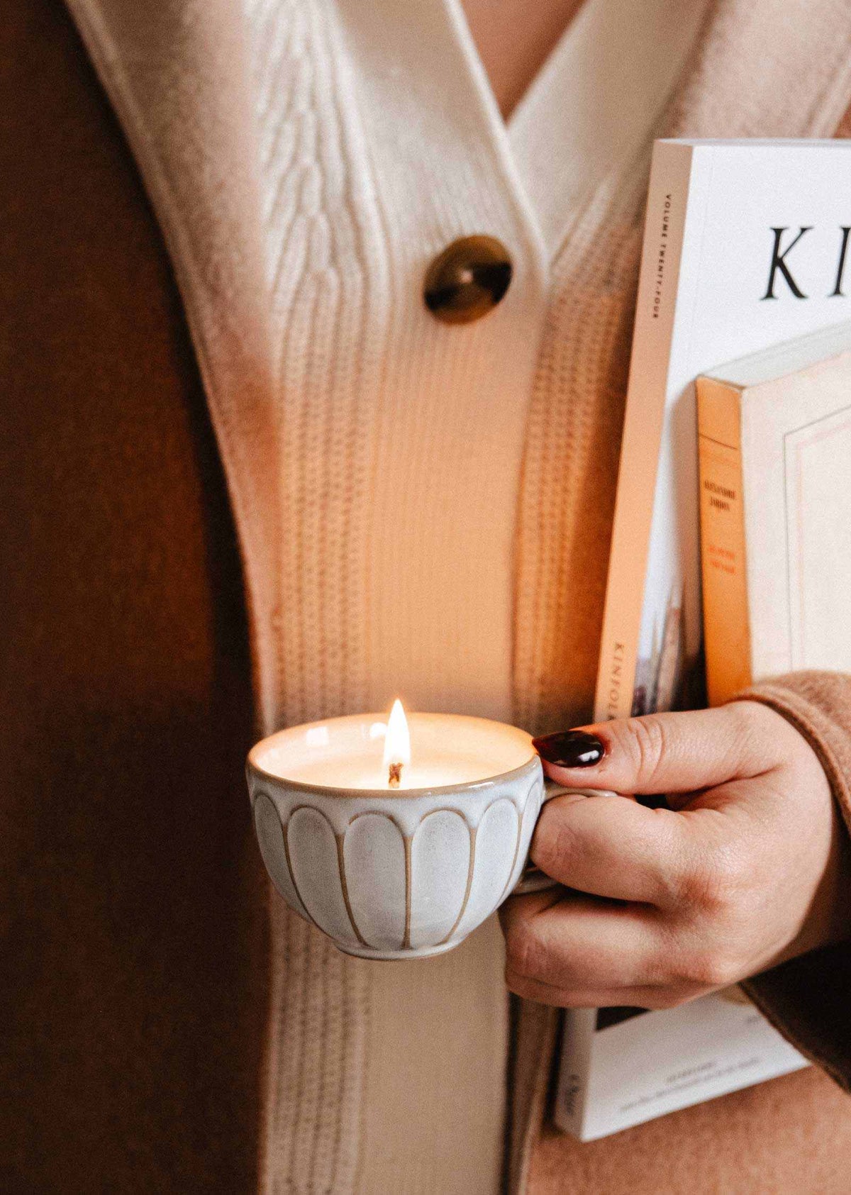 A person holds a lit Le Petit Café Mimi – Two Little Cups Candle Set in a small, fluted ceramic cup next to a stack of books.
