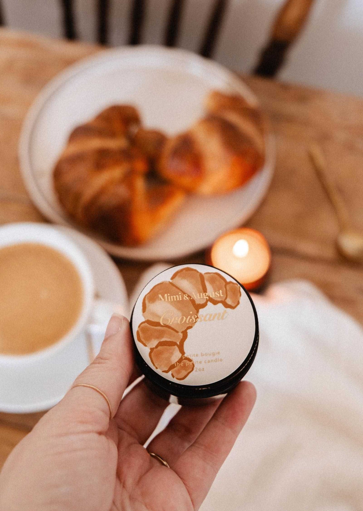 A hand holds Mimi & August’s Croissant – 2 oz Soy Candle in front of a table with a pastry, coffee, and a lit soy candle—invoking the cozy scent of a French bakery.