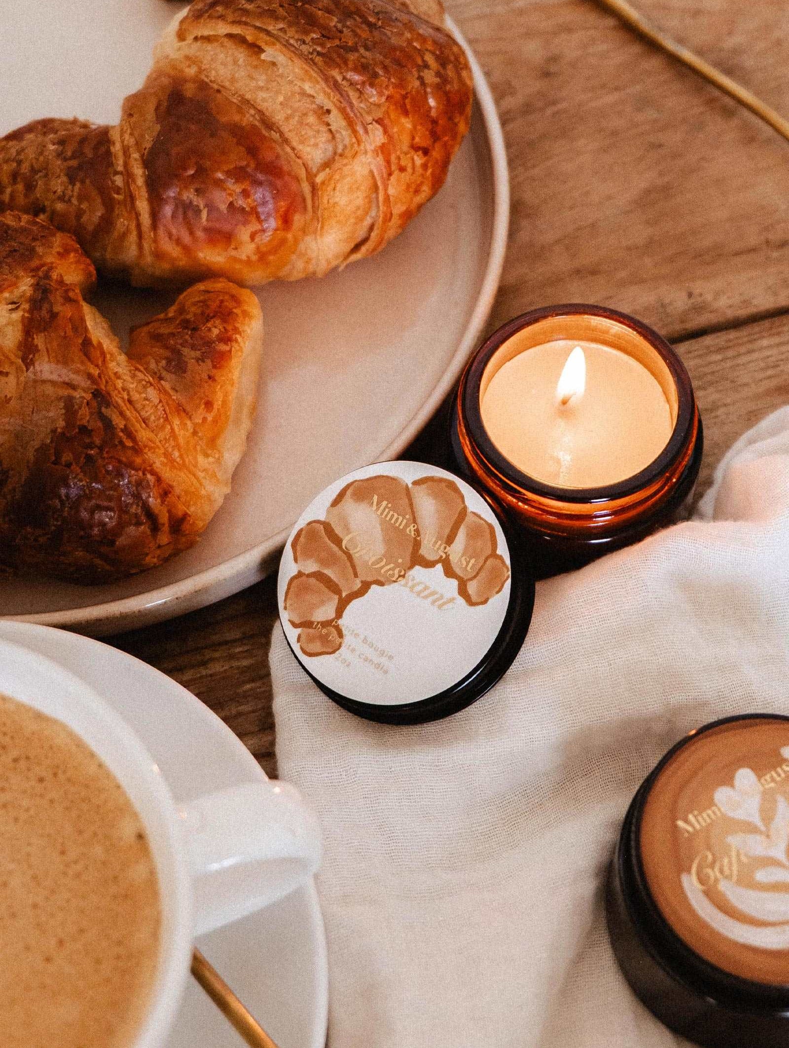 Une surface en bois accueille un croissant, un café et la bougie de soya Croissant de 2 oz de Mimi & August, évoquant le parfum réconfortant d'un matin de boulangerie française.