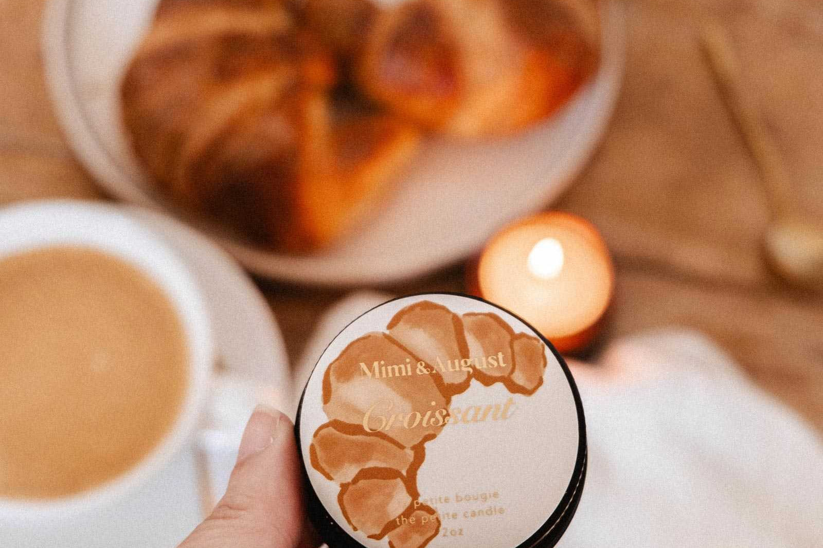 A hand holds Mimi & August’s Croissant – 2 oz Soy Candle in front of a table with a pastry, coffee, and a lit soy candle—invoking the cozy scent of a French bakery.