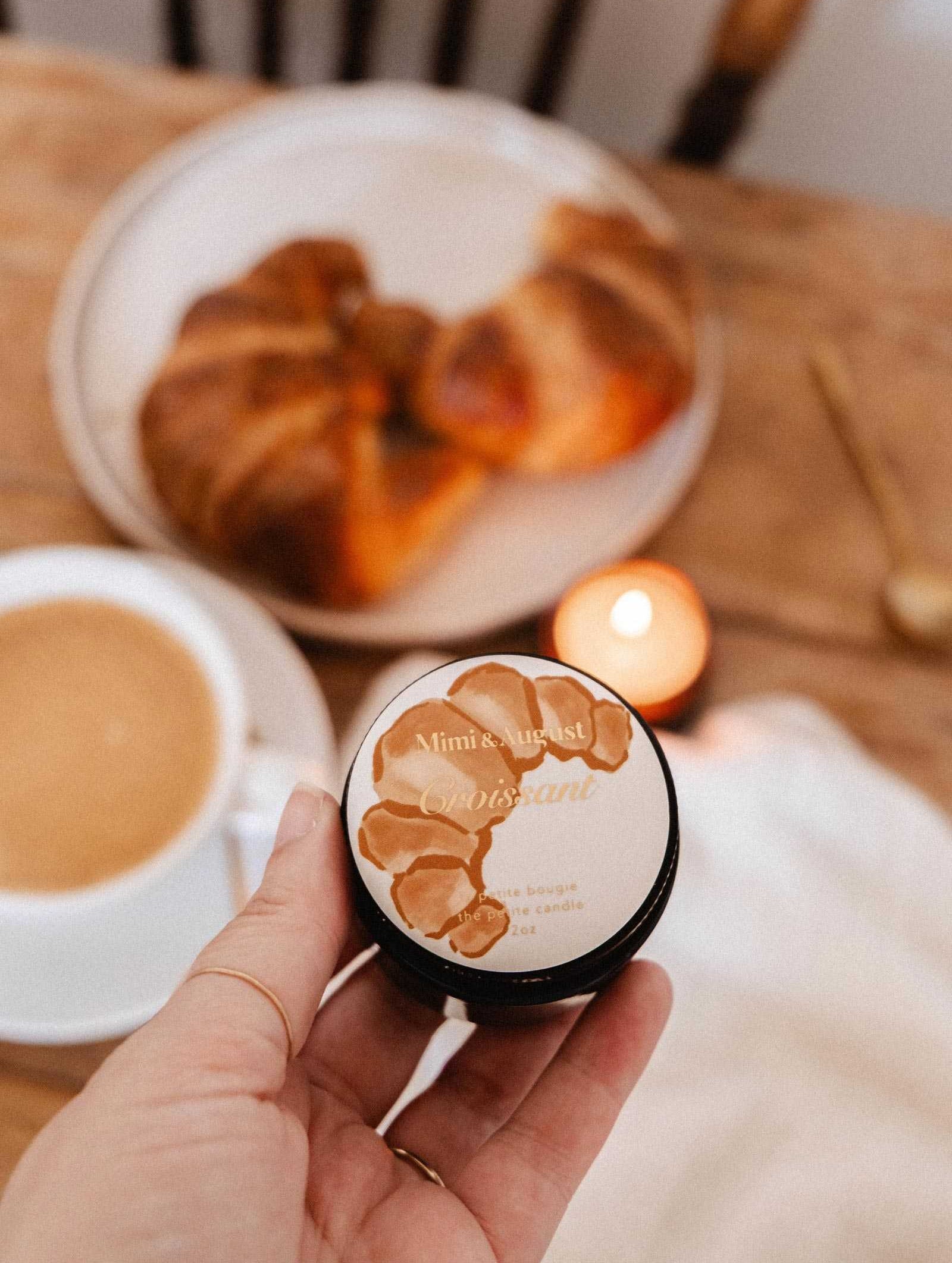 Une main tient la bougie de soya Croissant de 2 oz de Mimi & August devant une table avec une viennoiserie, un café et une bougie de soya allumée — évoquant le parfum réconfortant d'une boulangerie française.