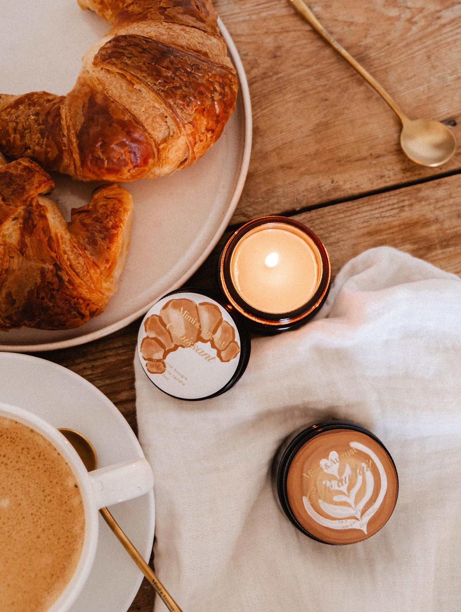 Une bougie de soya Croissant de 2 oz de Mimi & August brille à côté d'un vrai croissant, de deux petits pots, d'un café et d'une cuillère dorée, le tout disposé sur une table en bois avec un tissu blanc, évoquant l'arôme réconfortant d'une boulangerie française.