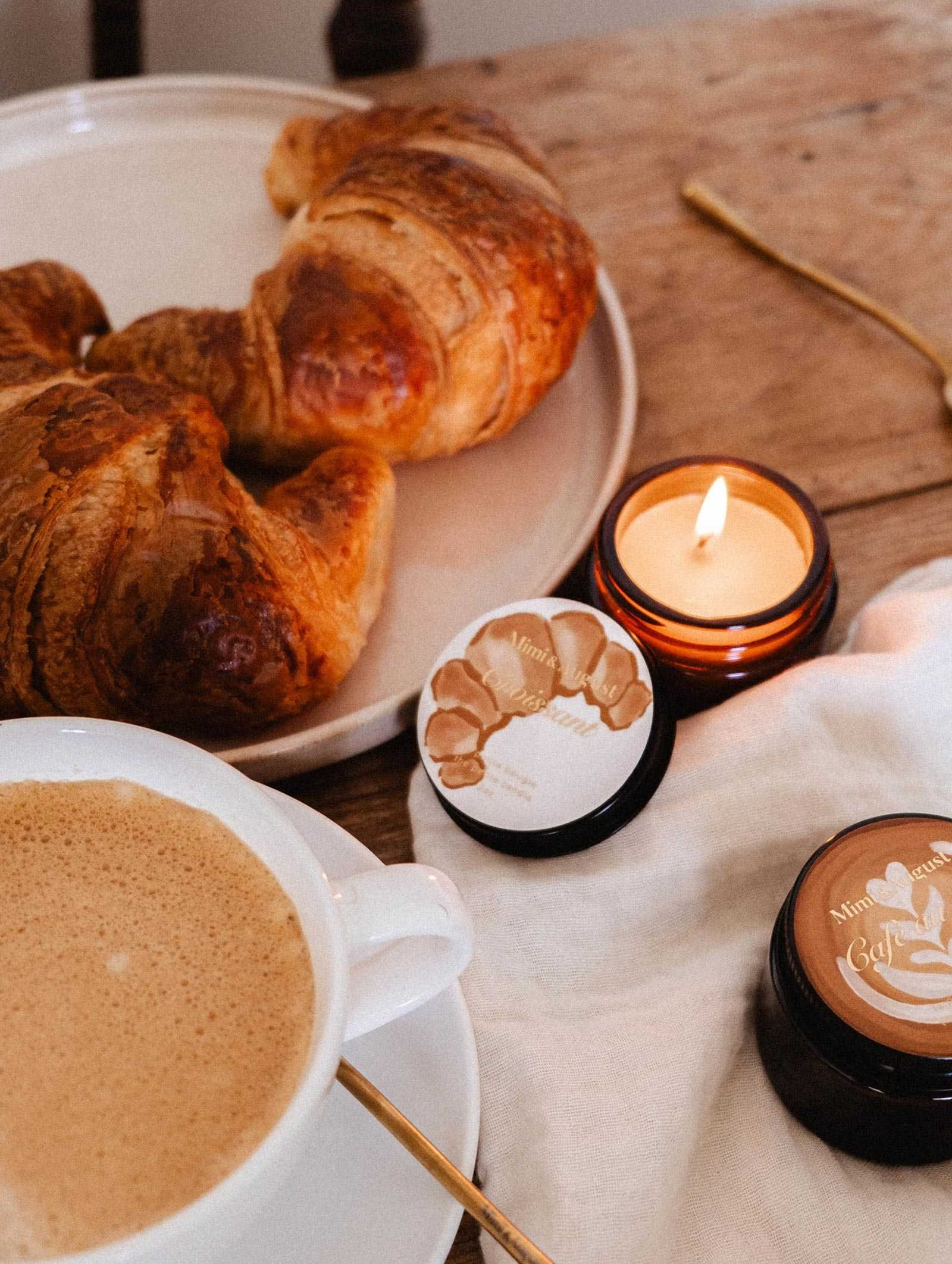 Une bougie de soya Croissant de 2 oz de Mimi & August repose sur une table en bois avec un tissu blanc, son subtil parfum de boulangerie française emplissant l'air à côté de deux croissants, d'une tasse de café et de deux pots de crème pour la peau.
