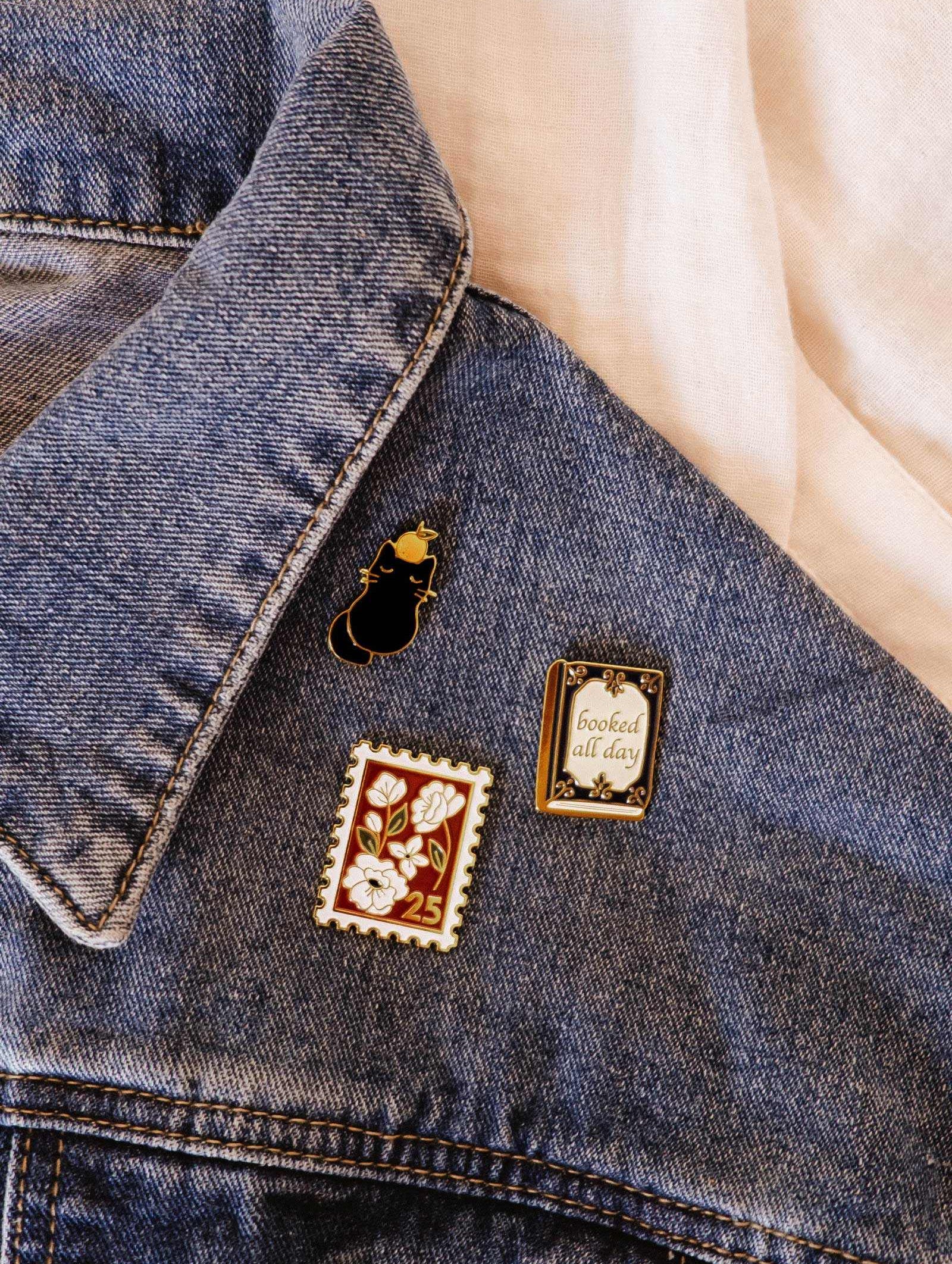 The collar of a blue denim jacket is adorned with three cute enamel pins, including the Clementine Cat Enamel Pin by Mimi & August, a floral “25” stamp, and a framed “booked all day” quote.