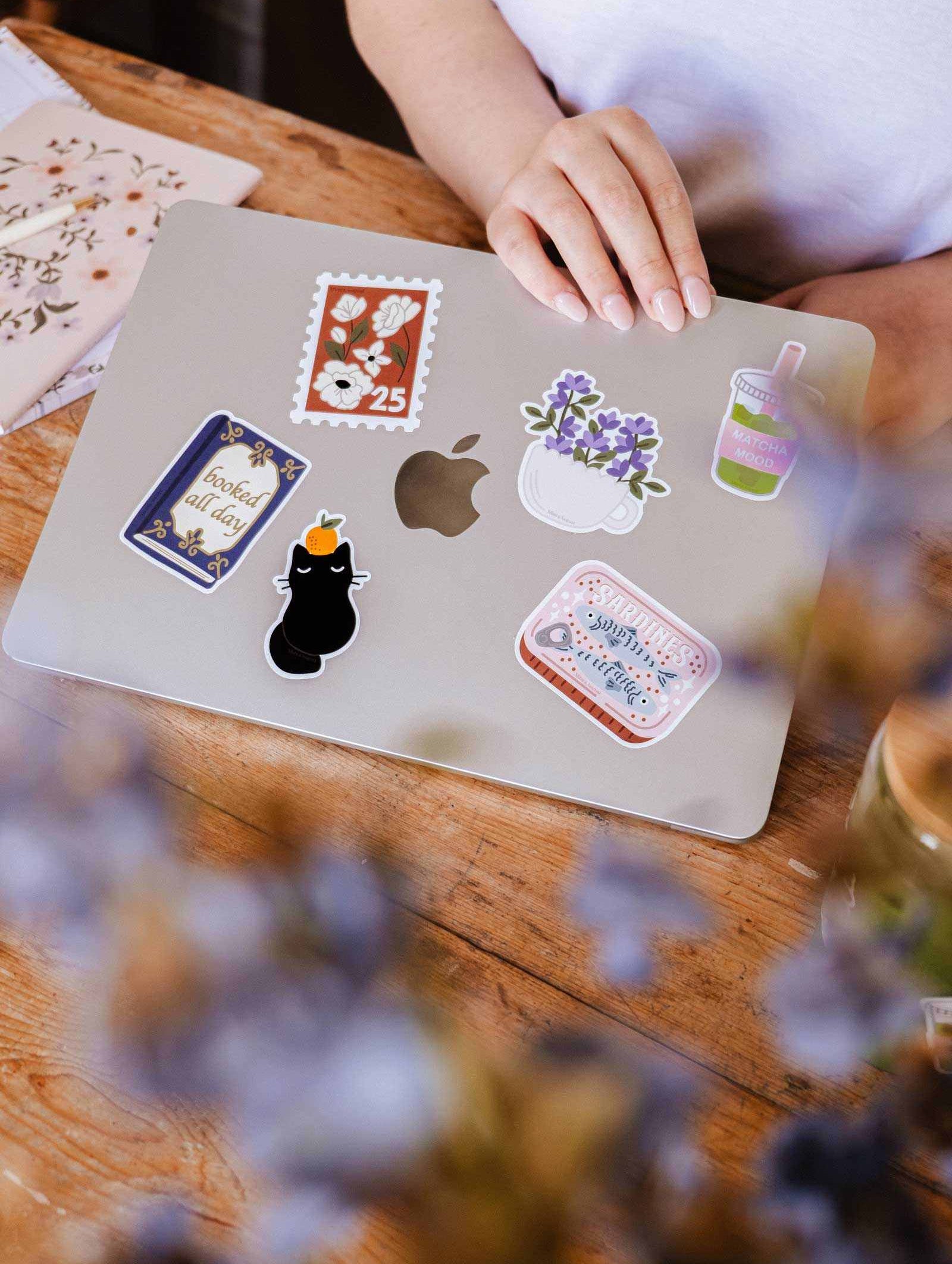 A hand rests on a silver MacBook adorned with colorful bookish stickers, including the Booked All Day - Vinyl Sticker by Mimi & August, on a wooden table with flowers and a reading journal sticker nearby.