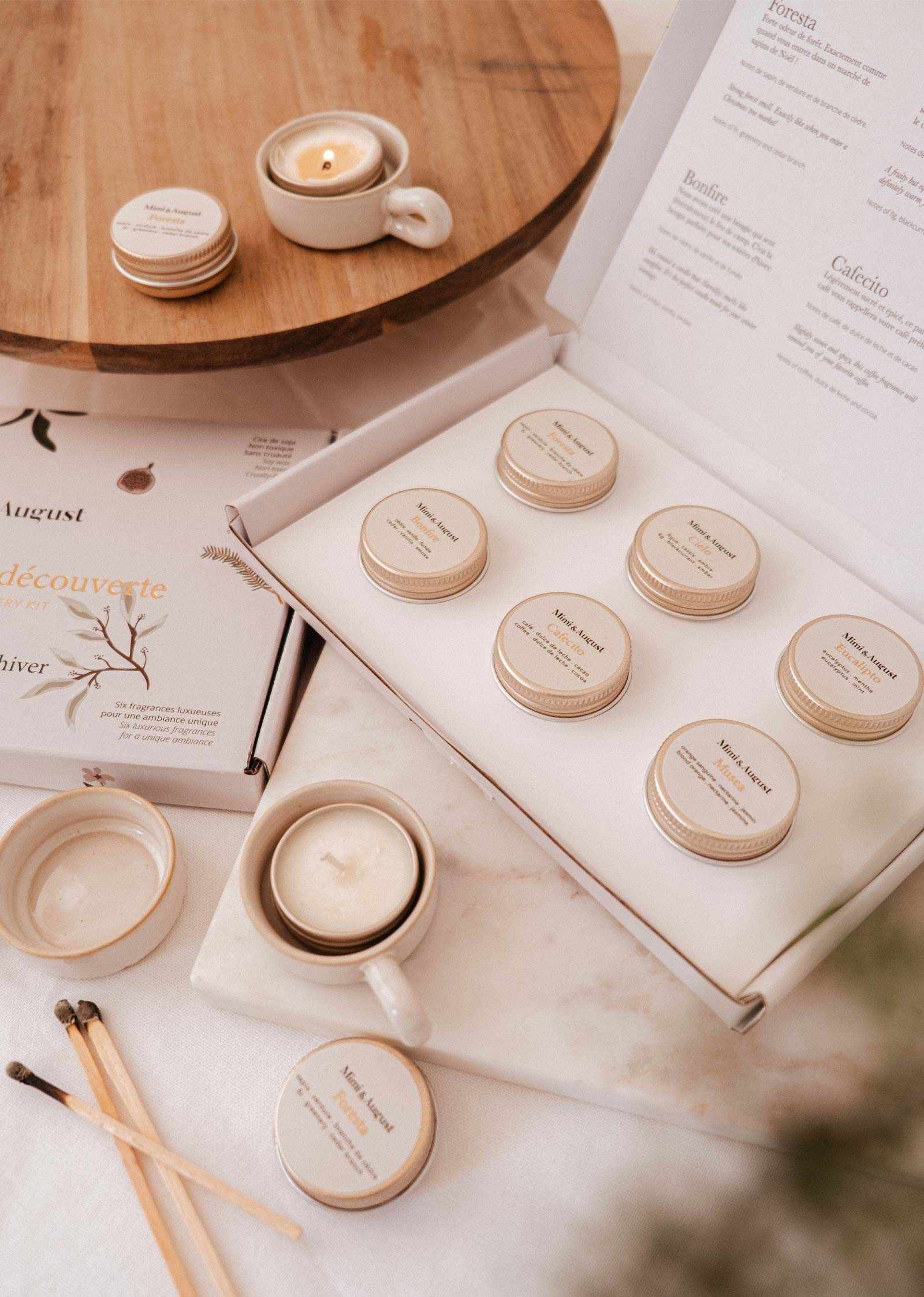 The Fall/Winter Discovery Kit by Mimi & August features small candle tins in an open white box with festive holiday scents, matches, and a lit coffee-scented candle nearby.