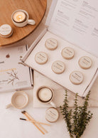 The Fall/Winter Discovery Kit by Mimi & August, featuring six small candles with beige lids, is open on a table beside cups, matches, greenery sprigs, and a candle holder—ideal for sampling holiday scents.