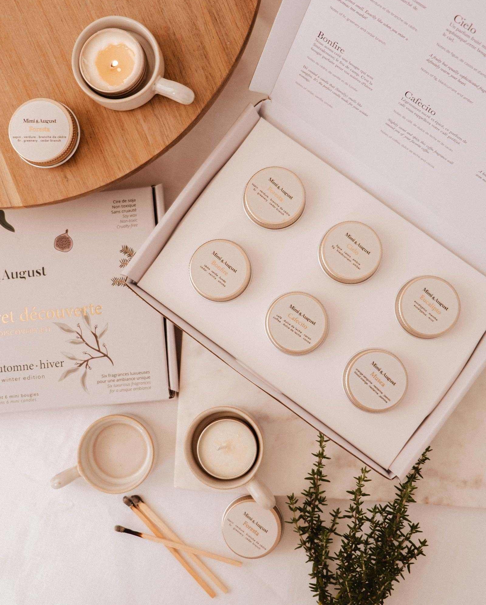 The Fall/Winter Discovery Kit by Mimi & August, featuring eight small holiday candle scents, is displayed beside a lit coffee candle, a cup, saucer, book, matches, and greenery on a light background.