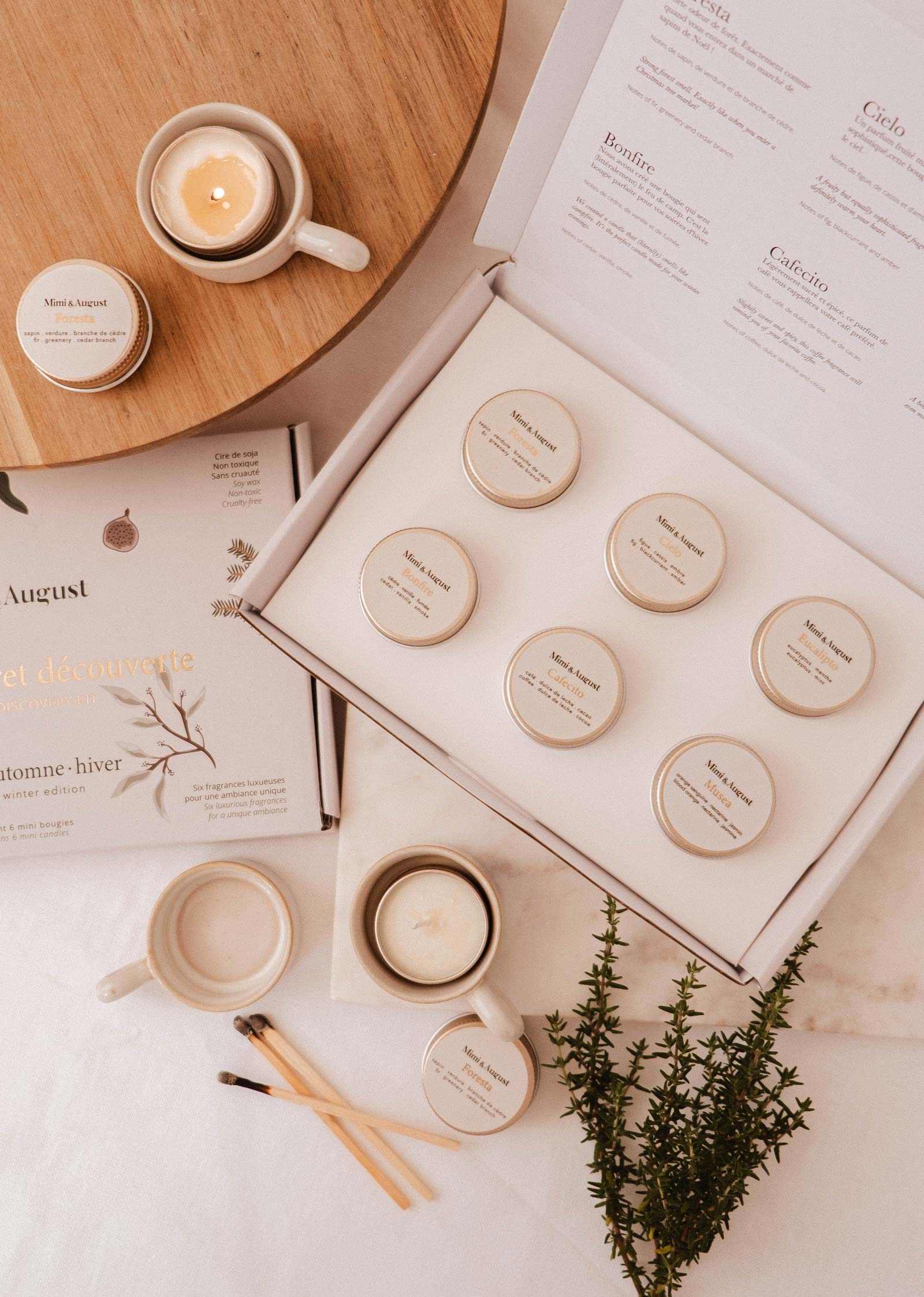 Coffret Bougies Automne/Hiver de Mimi & August, présentant huit petites bougies aux parfums des fêtes, est exposé à côté d'une bougie au café allumée, d'une tasse, d'une soucoupe, d'un livre, d'allumettes et de verdure sur un fond clair.