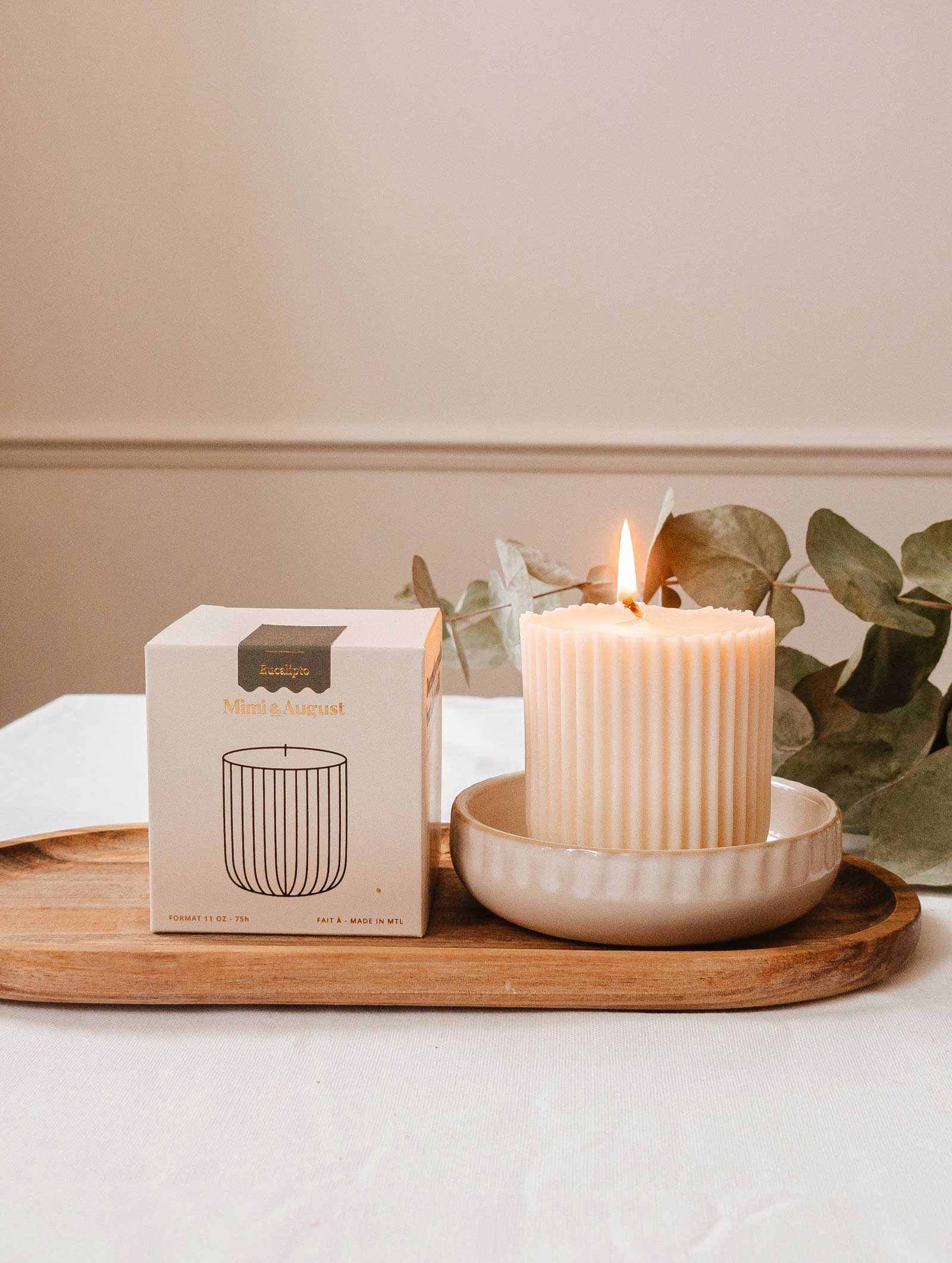 La Recharge de bougie Eucalipto de Mimi & August brille dans un plat en céramique sur un plateau en bois près de sa boîte, avec des branches d'eucalyptus en arrière-plan.