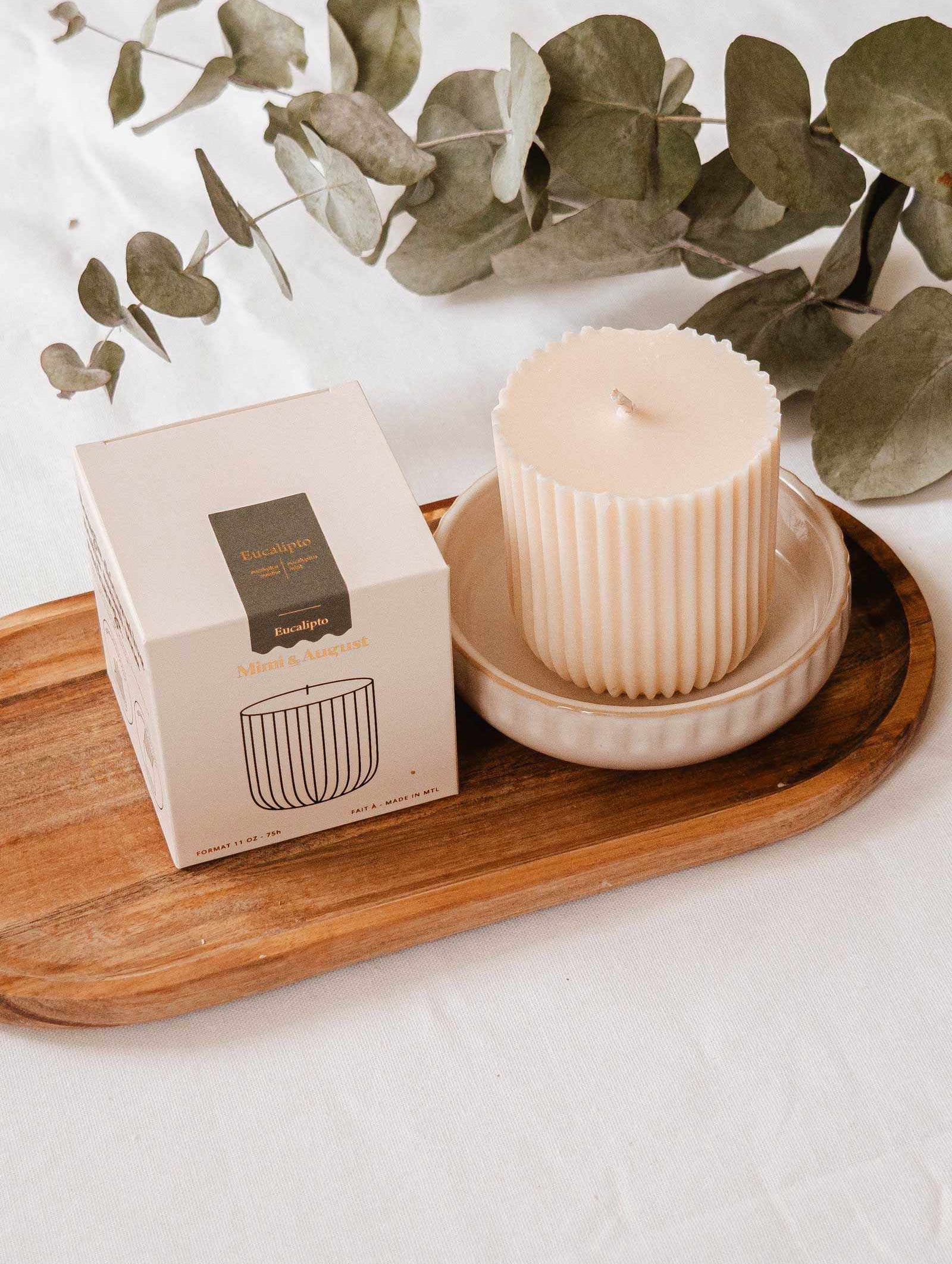 Une bougie écoresponsable cannelée de couleur crème dans un plat en céramique repose sur un plateau en bois à côté de sa boîte, avec des feuilles d'eucalyptus derrière — à associer parfaitement avec la Recharge de bougie Eucalipto de Mimi & August.