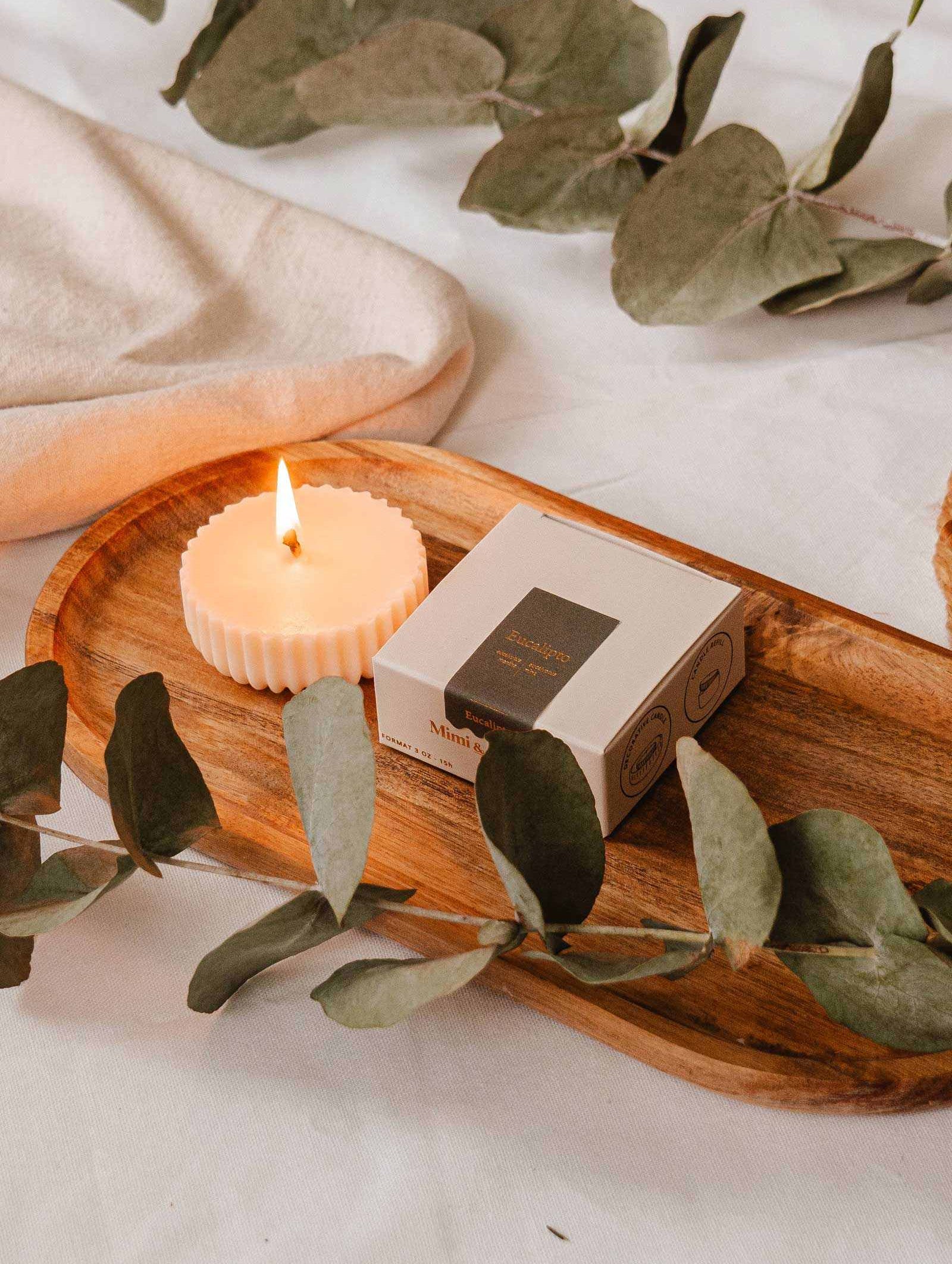 La Recharge de bougie Eucalipto de Mimi & August repose élégamment sur un plateau en bois avec une boîte, entourée de feuilles d'eucalyptus luxuriantes sur une surface blanche, offrant une touche écoresponsable.