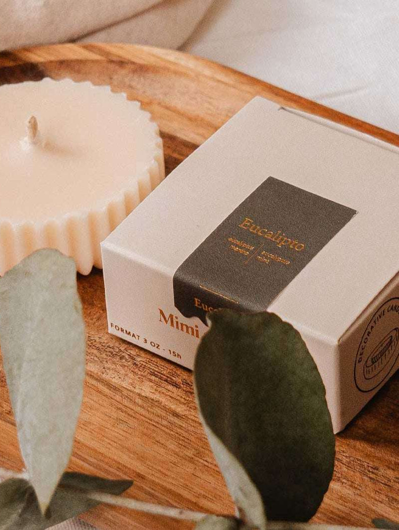Une Recharge de bougie Eucalipto de Mimi & August et un article en boîte reposent sur un plateau en bois, entourés de feuilles d'eucalyptus sur une surface blanche, créant une pièce maîtresse écoresponsable.