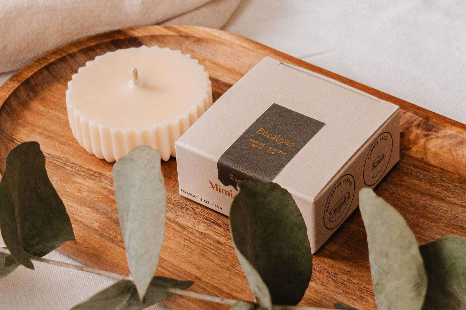 Une Recharge de bougie Eucalipto de Mimi & August et un article en boîte reposent sur un plateau en bois, entourés de feuilles d'eucalyptus sur une surface blanche, créant une pièce maîtresse écoresponsable.