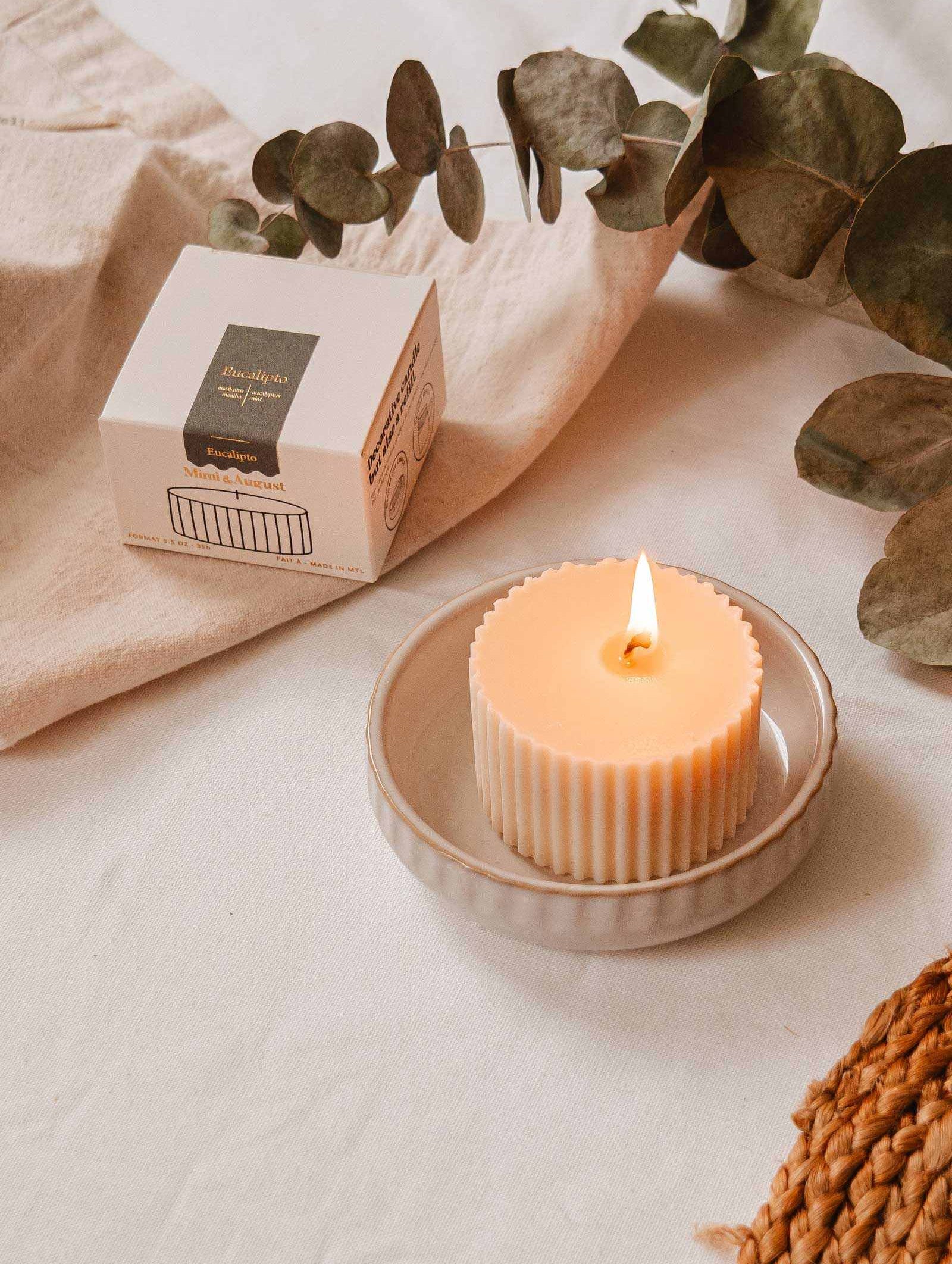La Recharge de bougie Eucalipto de Mimi & August repose sur une petite assiette à côté de sa boîte sur un tissu blanc, agrémentée de feuilles d'eucalyptus et d'un napperon tressé.