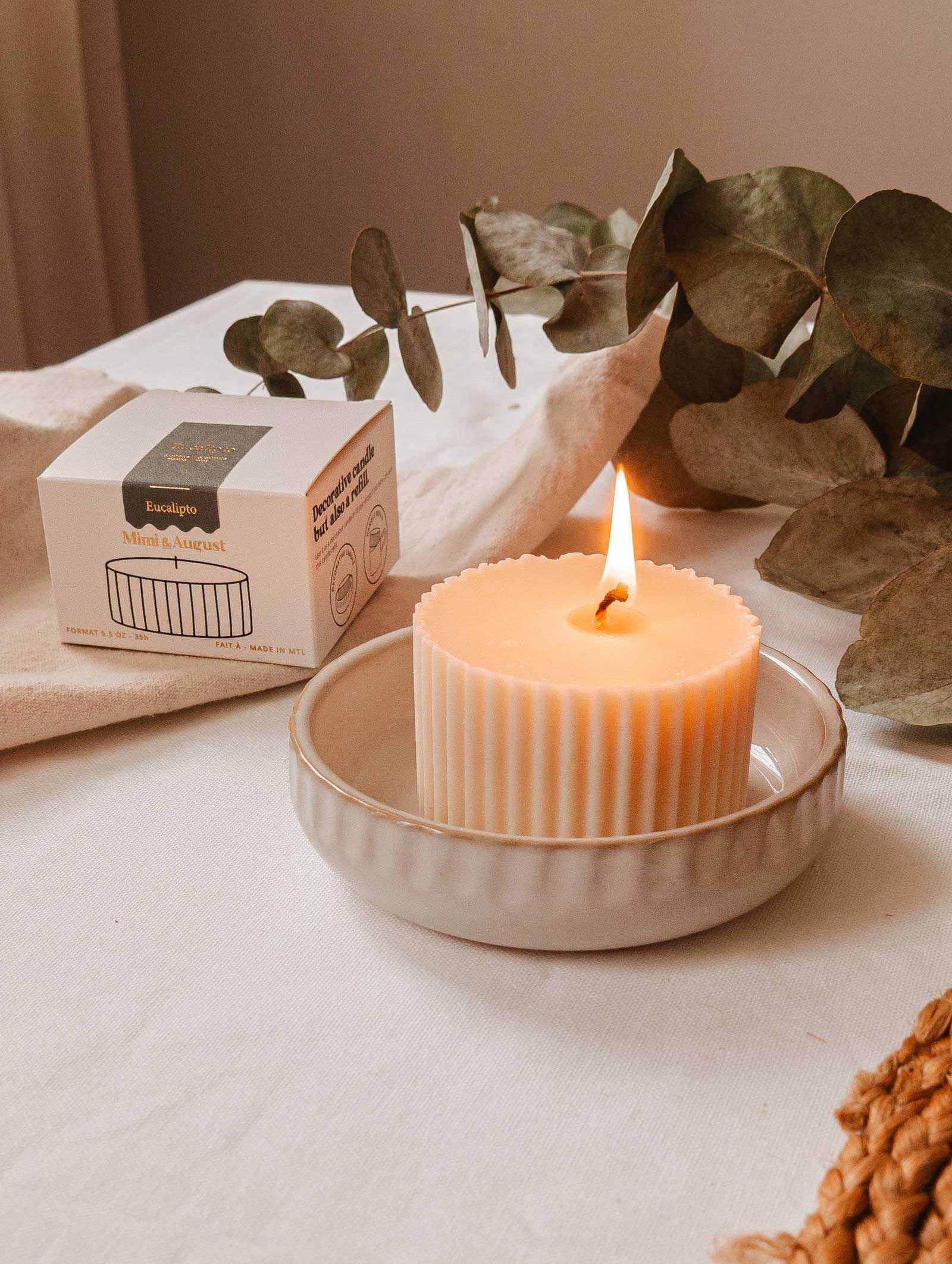 La Recharge de bougie Eucalipto de Mimi & August, gracieusement allumée dans un plat en céramique aux côtés de sa boîte écoresponsable et de feuilles d'eucalyptus luxuriantes, élégamment disposées sur une surface recouverte de tissu.