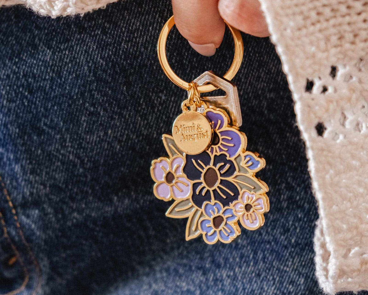 Une main tient le Porte-clés Bouquet Fleuri de Mimi & August et deux clés, sur un fond de jeans en denim bleu et un chandail en crochet crème.