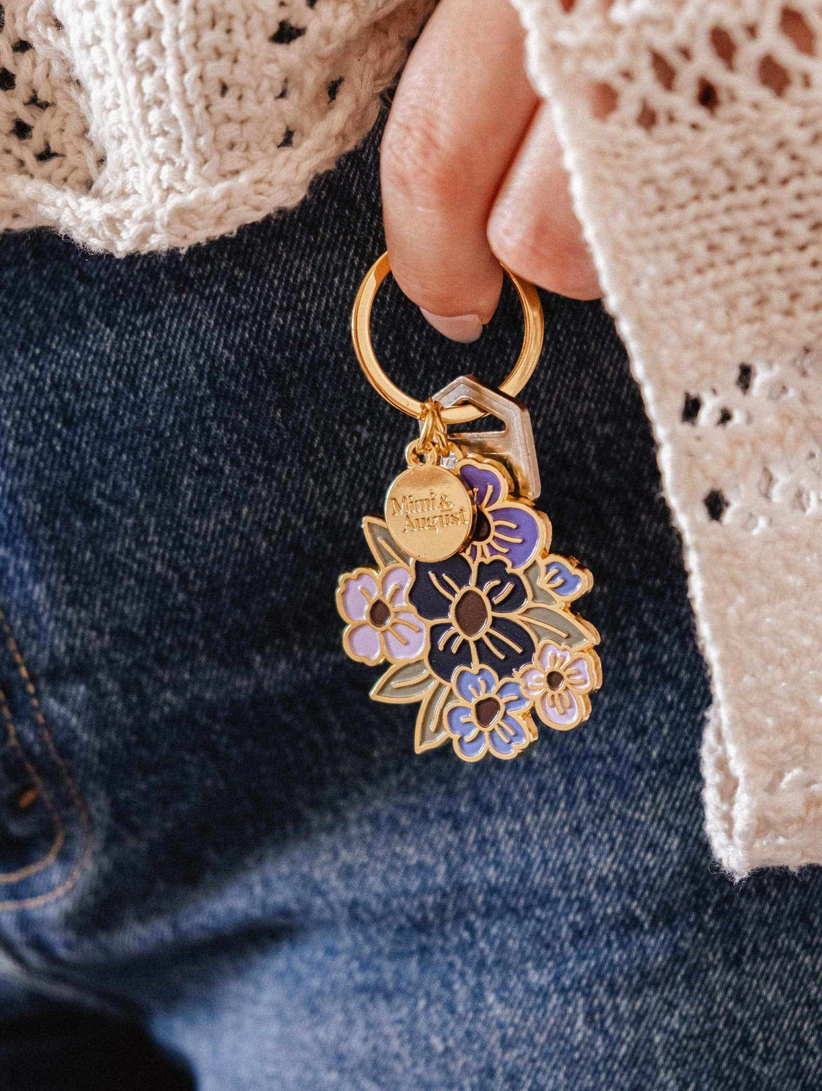 Une main tient le Porte-clés Bouquet Fleuri de Mimi & August et deux clés, sur un fond de jeans en denim bleu et un chandail en crochet crème.