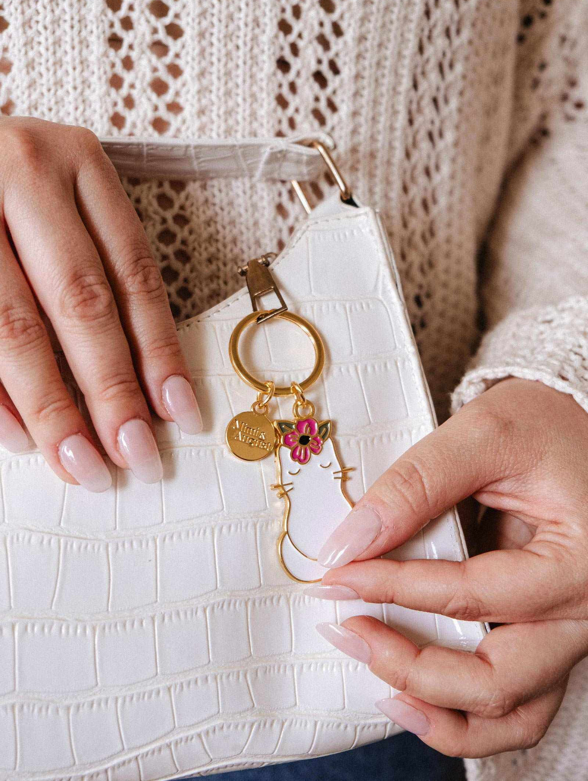 Une personne aux ongles manucurés tient un sac à main blanc texturé avec le Porte-clés Chat Fleuri de Mimi & August, en forme de chat avec une fleur rose — un accessoire idéal pour les amoureux des chats.